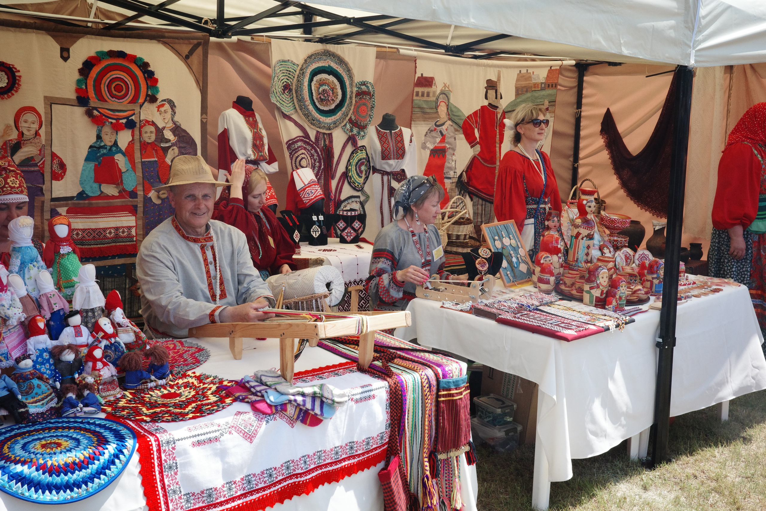 Мастер за ткацким станком, рядом народные куклы и вышитые изделия в сувенирном шатре