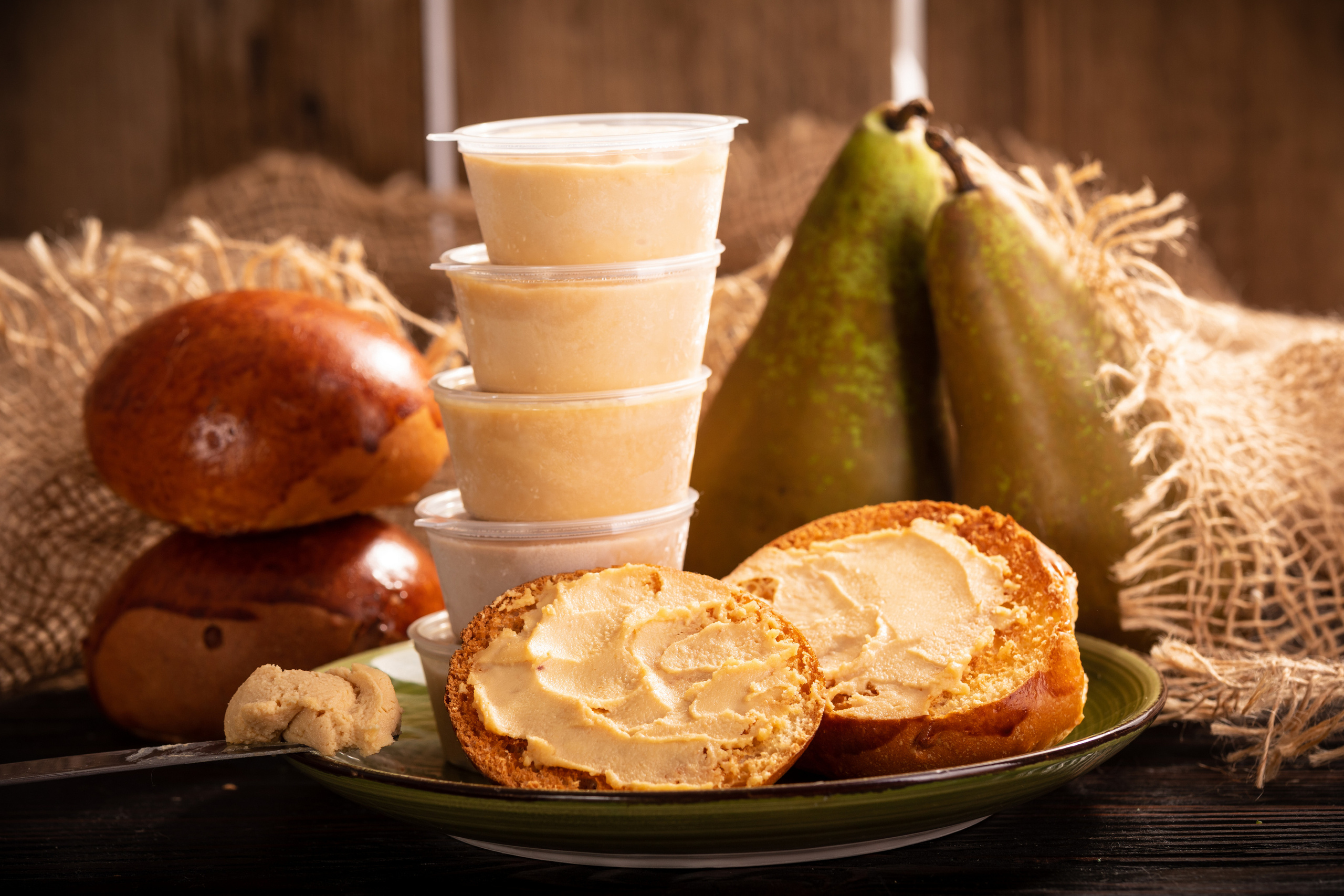 Два бутерброда с сыром брюност и половинка зеленой груши на тарелке. Рядом чашка кофе, две груши и зелень гороха на столе из дерева.