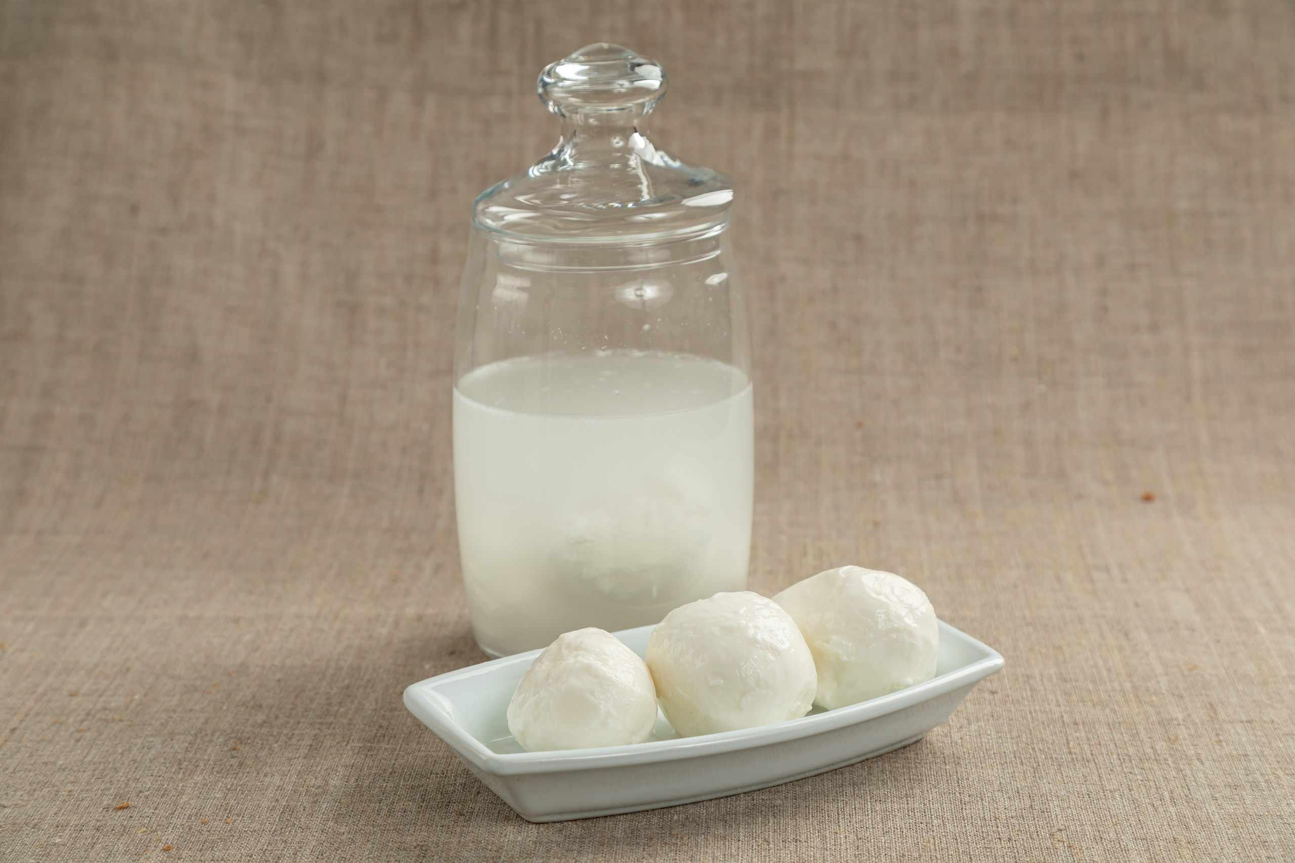 Три больших шарика моцареллы в белой прямоугольной тарелке. На заднем плане стеклянная банка с рассолом и шариком сыра.