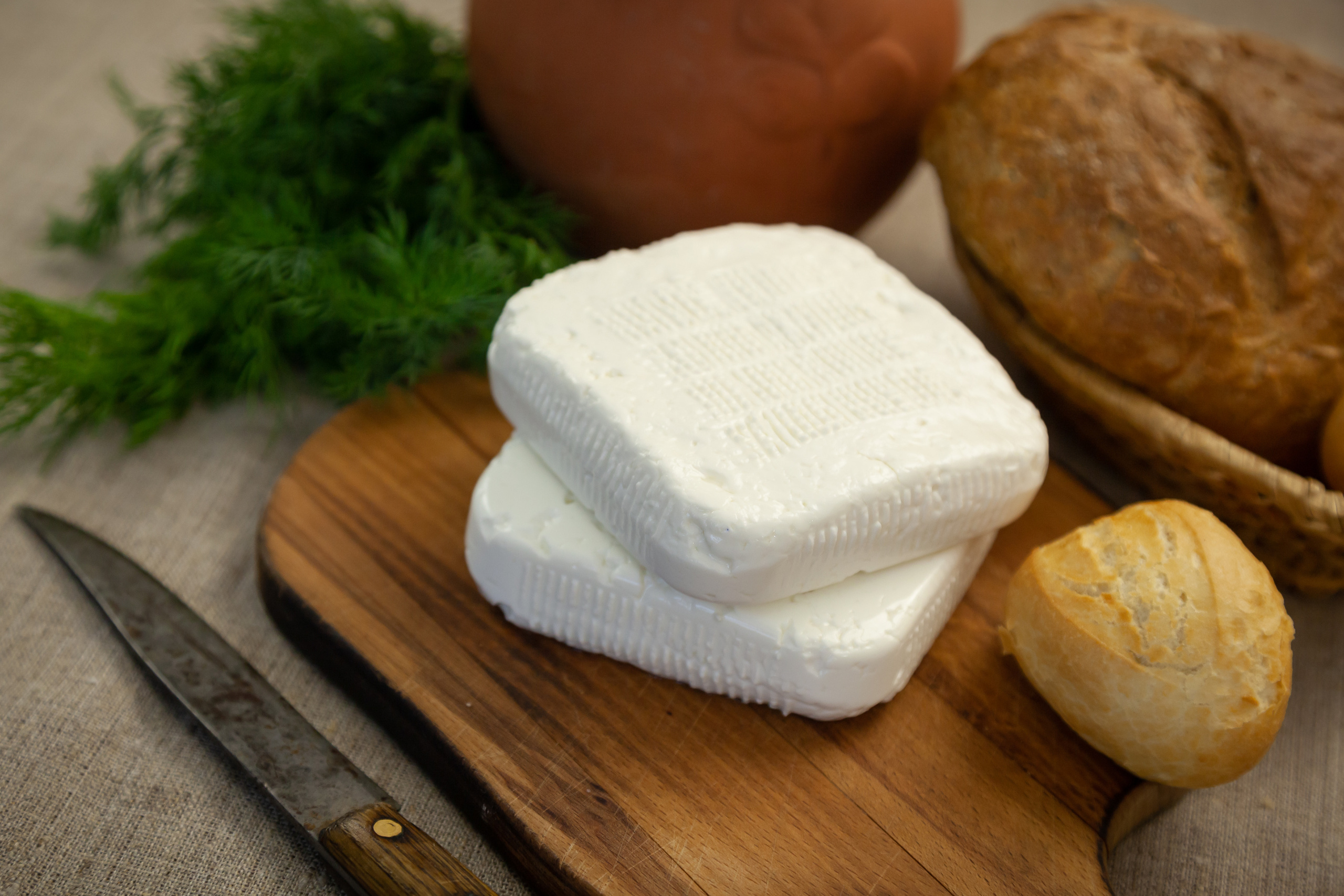 две головки имеретинского сыра на досочке. Рядом нож, хлеб, укроп и глиняный кувшин.