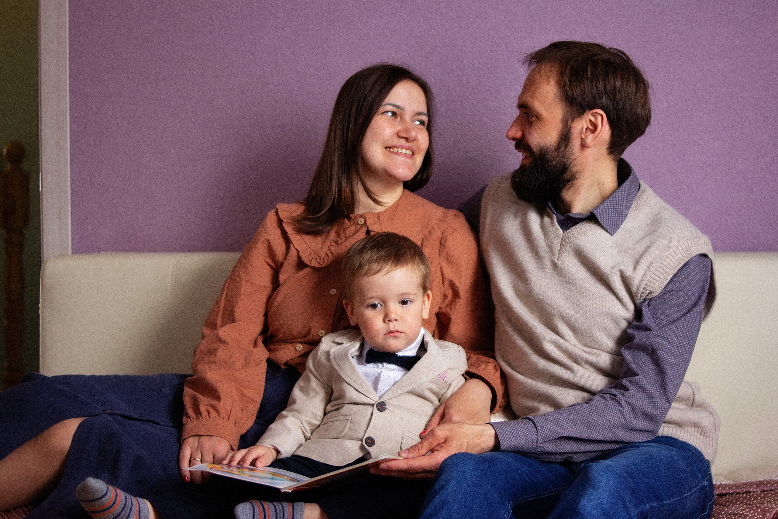 муж и жена смотрят друг другу в глаза и счастливо улыбаются между ними сидит их двух летний сын и смотрит книгу