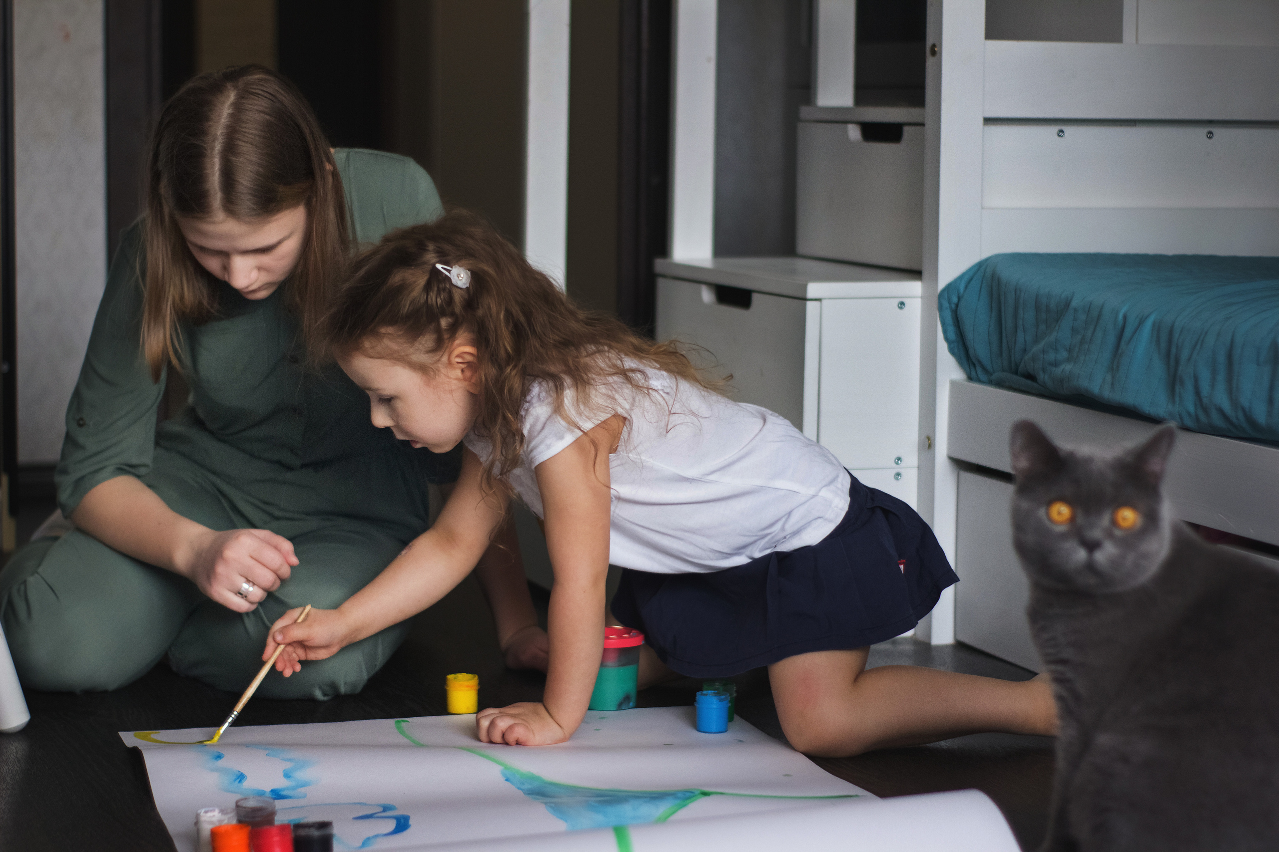 Две девочки сестренки рисуют дома красками на большом ватмане рядом сидит красивый серый кот с чайными глазами