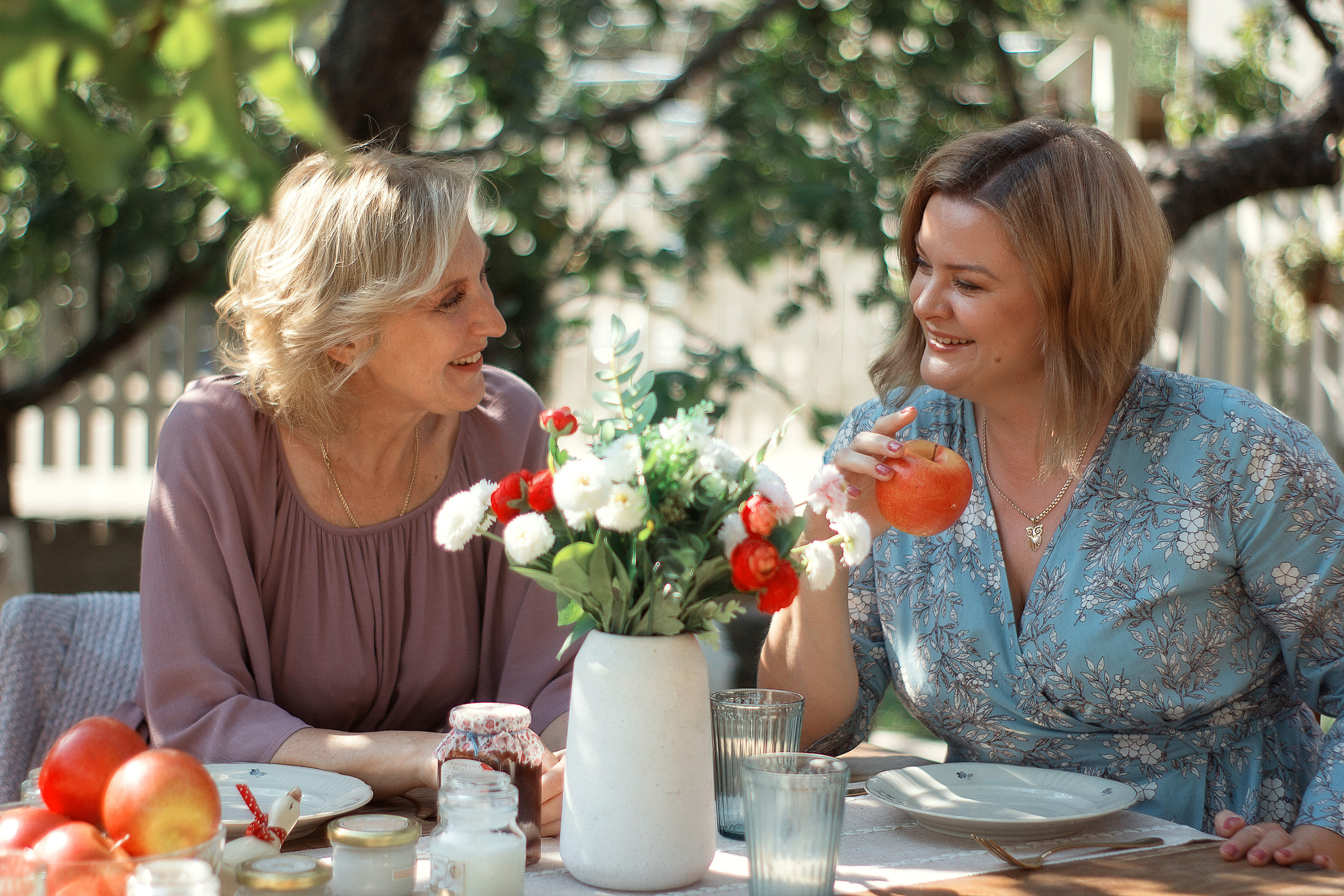 Две девушки разговаривают и улыбаются сидя за изящно накрытым столом. Разговор подруг за чаепитием под яблоней.