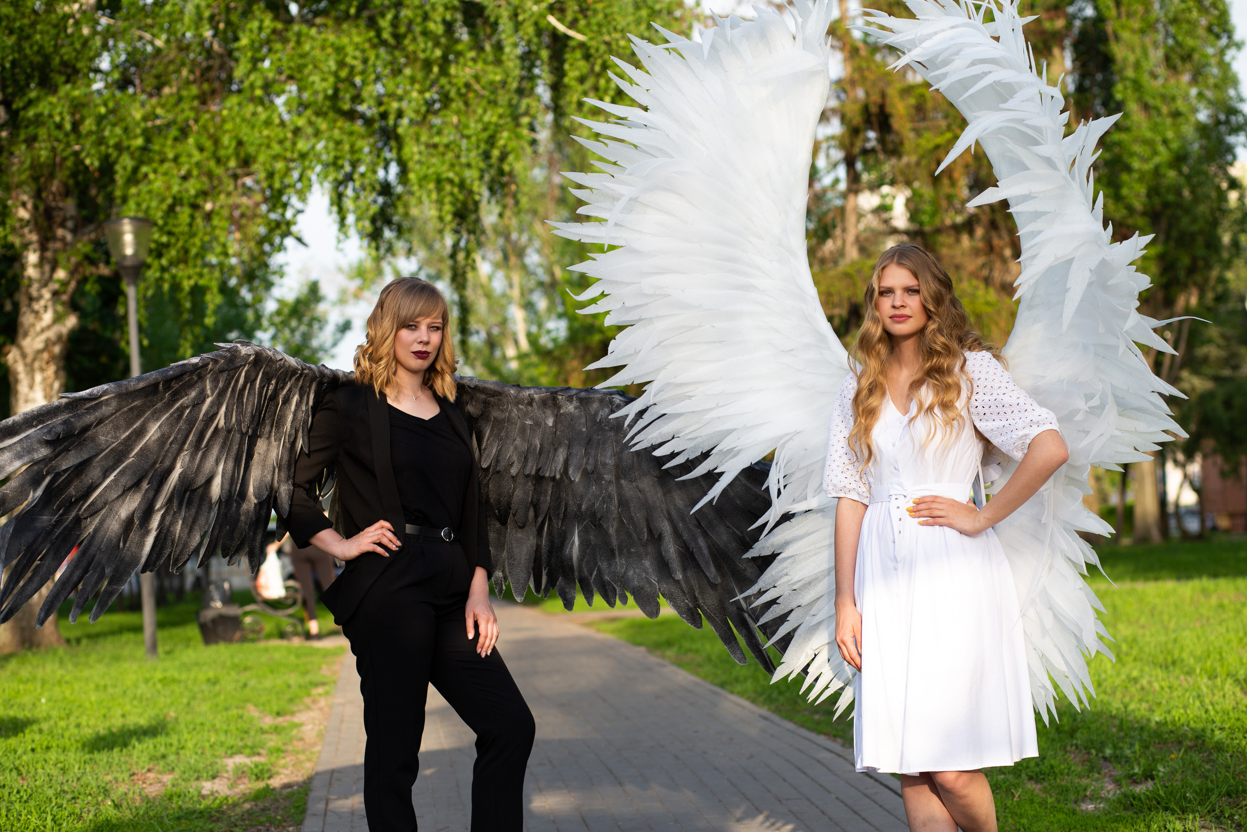 две девушки. одна  в черном и у нее черные  крылья. вторая   в белом  платье и у нее белые крылья. стоят в парке. прокат крыльев для фотосессий Воронеж. изготовление крыльев для мероприятий  и фото сессий на  заказ. 
