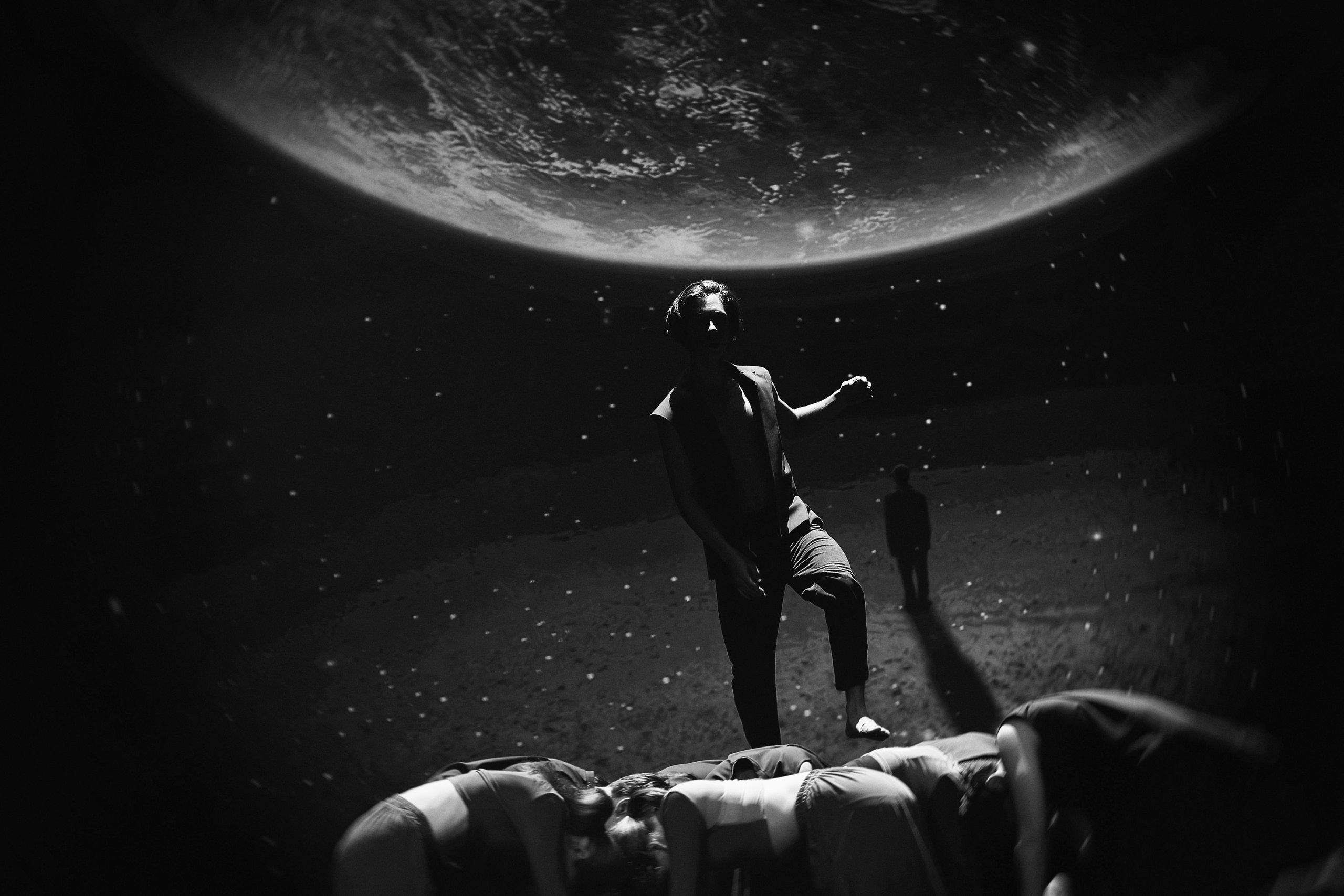 Танцор стоит под проекцией планеты Земля со звездами, другие танцоры лежат на полу, космическая тематика