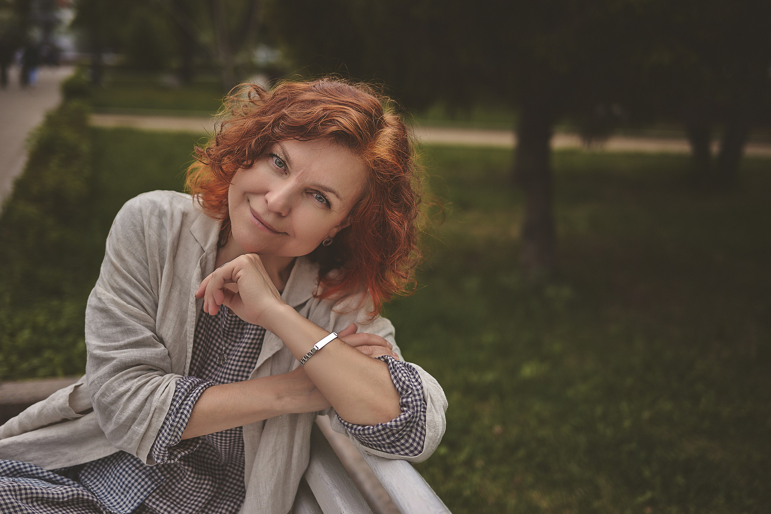 Женщина с вьющимися рыжими волосами, мягко улыбающаяся, сидит на скамейке на открытом воздухе, одетая в  свободный льняной пиджак поверх клетчатого платья. На заднем плане изображена пышная зелень.
