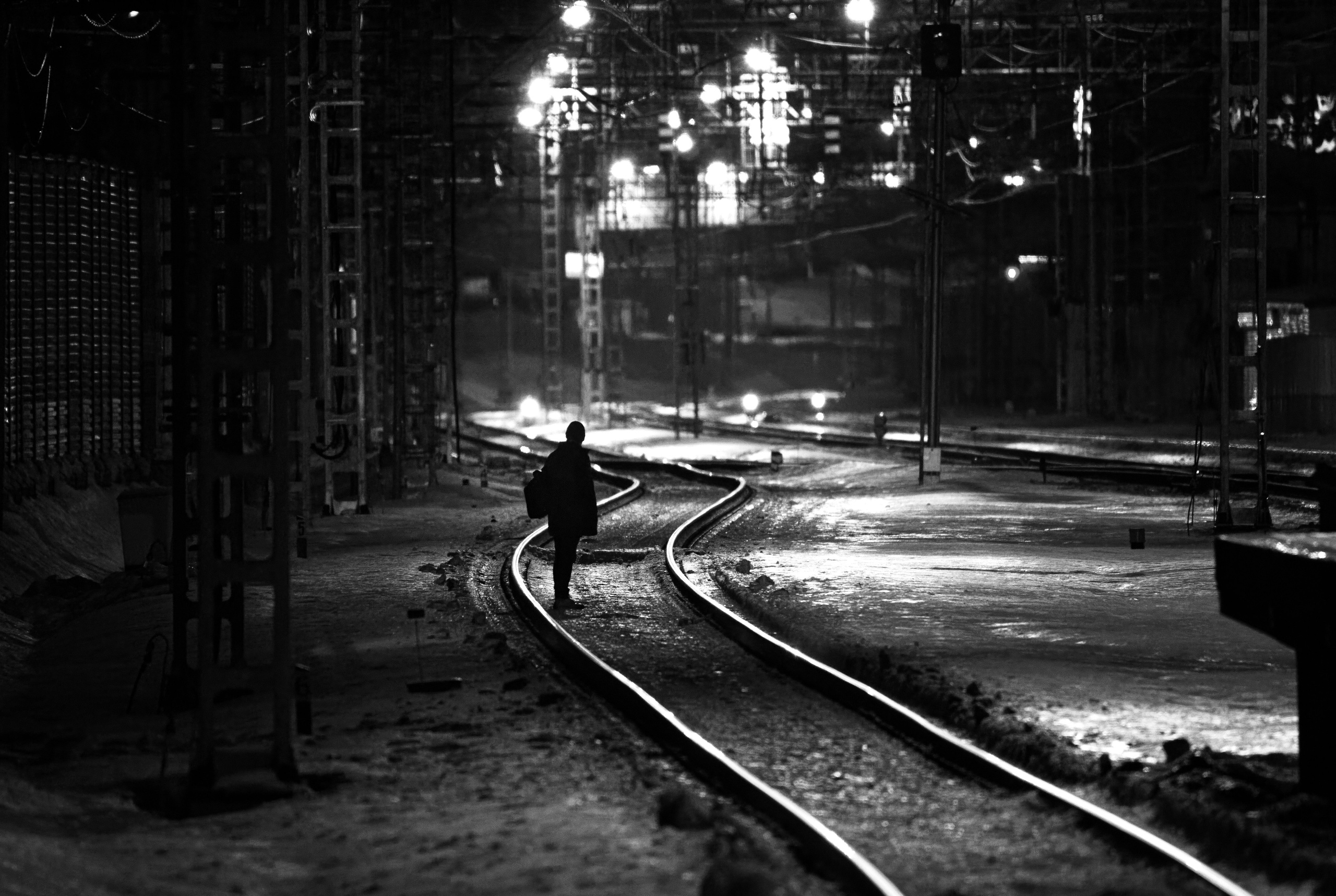 Черно-белое ночное фото человека идущего по железнодорожным путям, огни города на фоне, силуэт, урбанистическая атмосфера