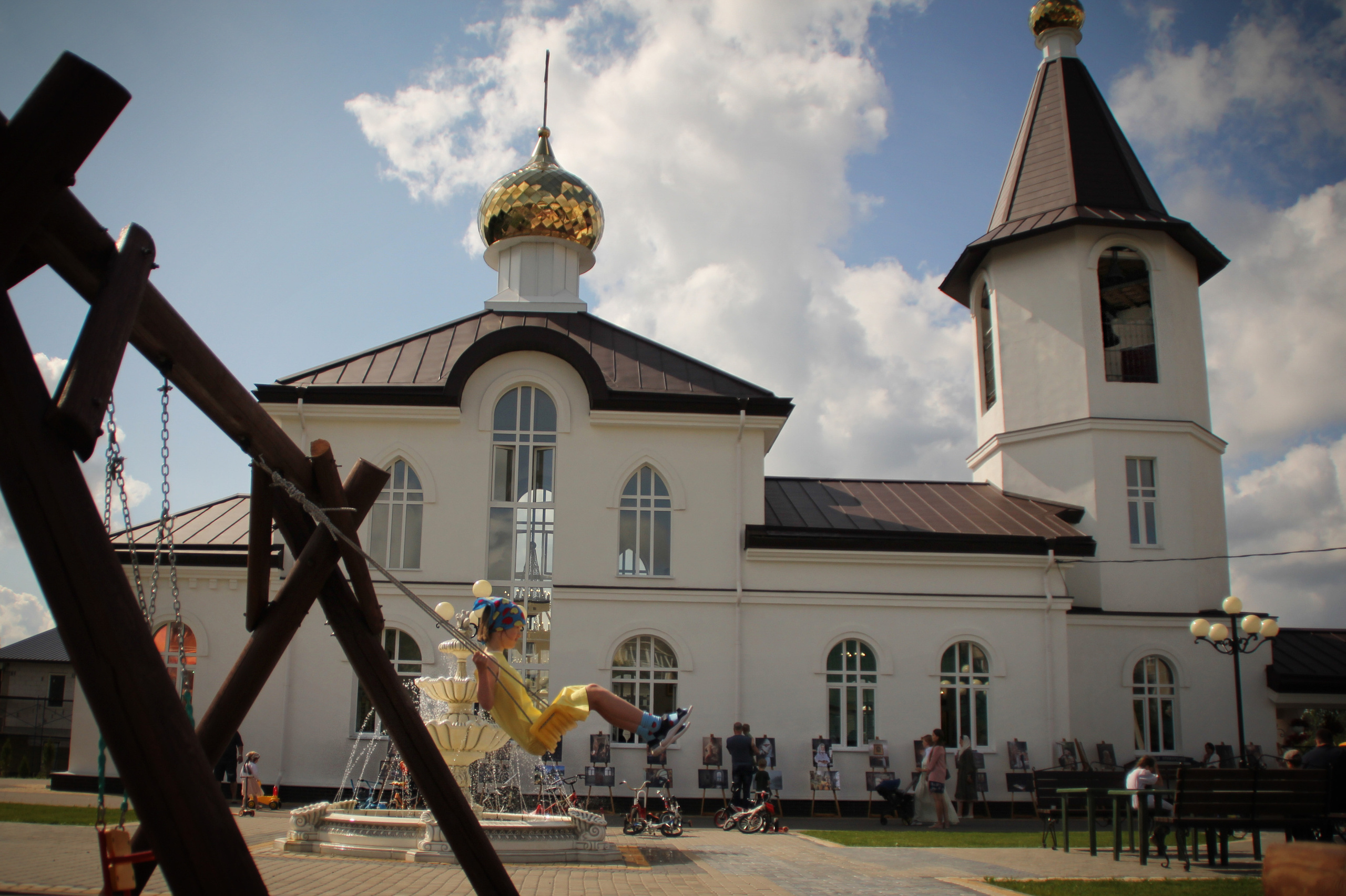 девочка в желтом платье качается на качелях во дворе храма на фоне голубого неба и золотых куполов