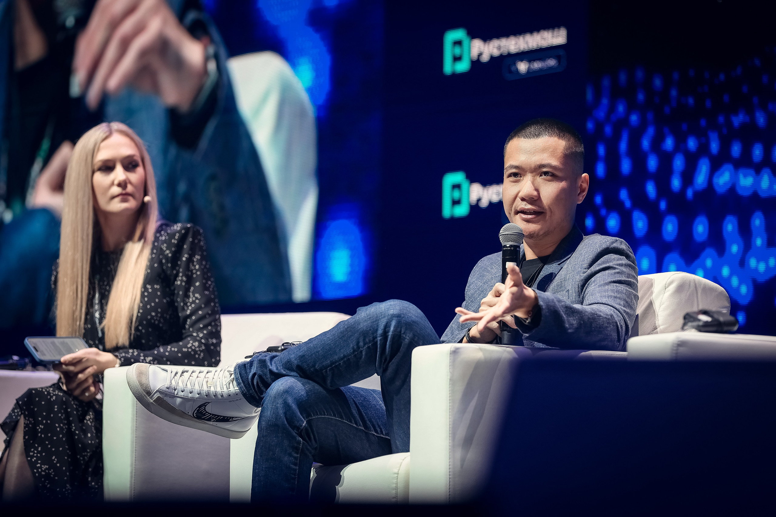 Средний план: представитель Бинг Гиггс продолжает выступление, девушка-спикер слушает внимательно, нога на ногу, спокойно и сосредоточенно.