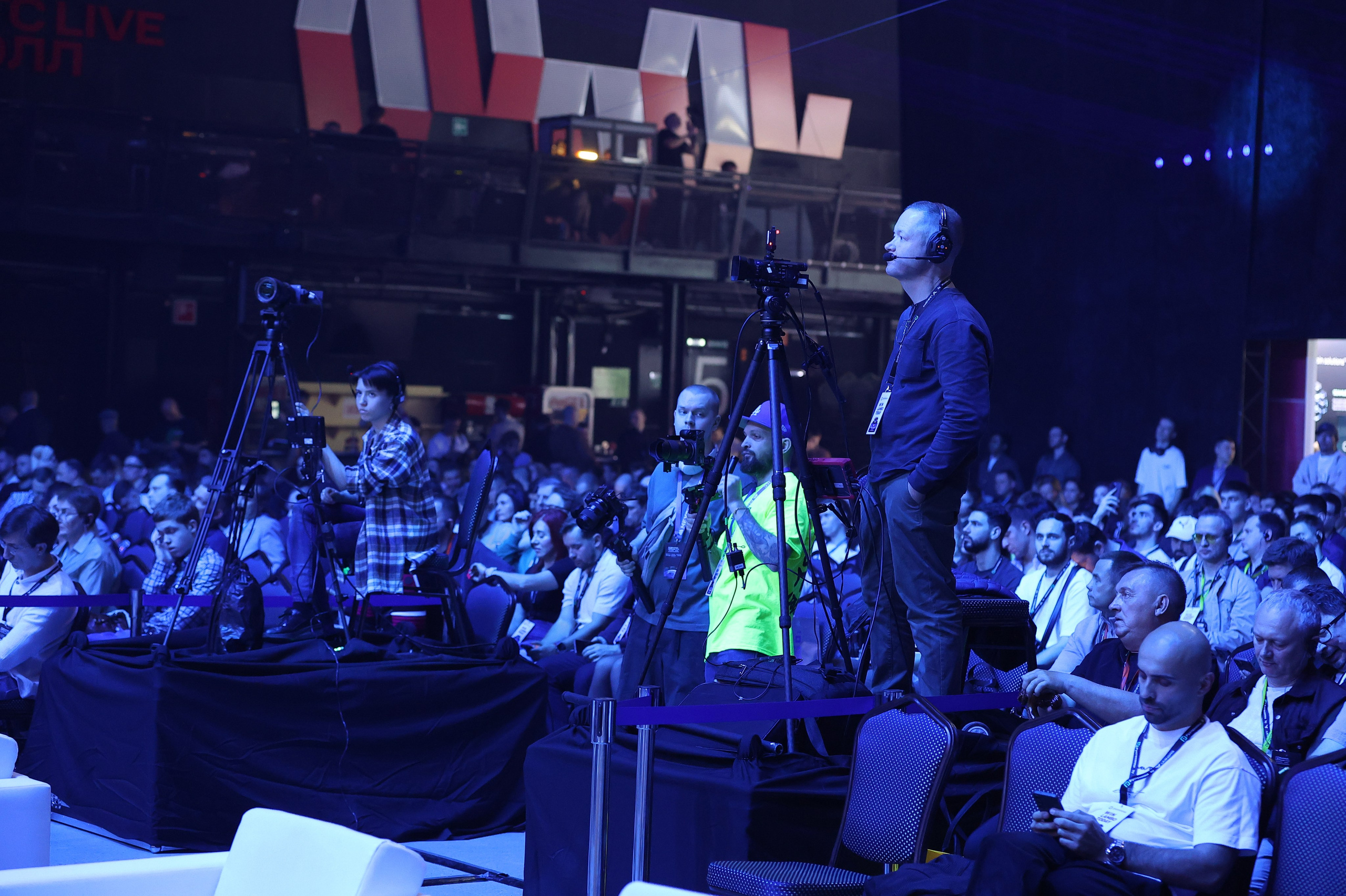 Видеооператор в наушниках наблюдает за съемкой и зрителями, стоит на месте, несколько операторов вокруг, один в фокусе.