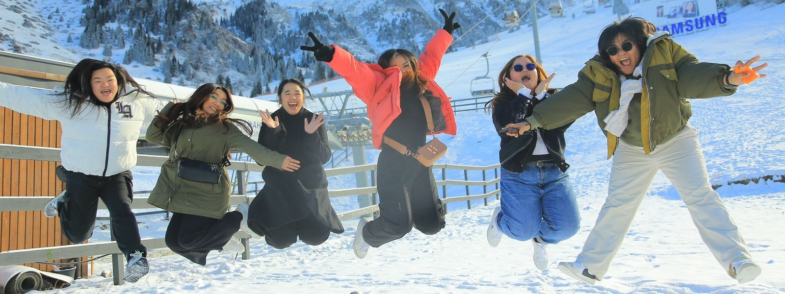 Группа из шести человек в зимней одежде прыгает на фоне заснеженных гор. Все выглядят счастливыми и веселыми, наслаждаясь снежной атмосферой и солнечным днем. Вдали виднеются горные склоны с густыми лесами, а также канатная дорога Шымбулак. Небо ясное с небольшими облаками, создавая живописный зимний пейзаж.