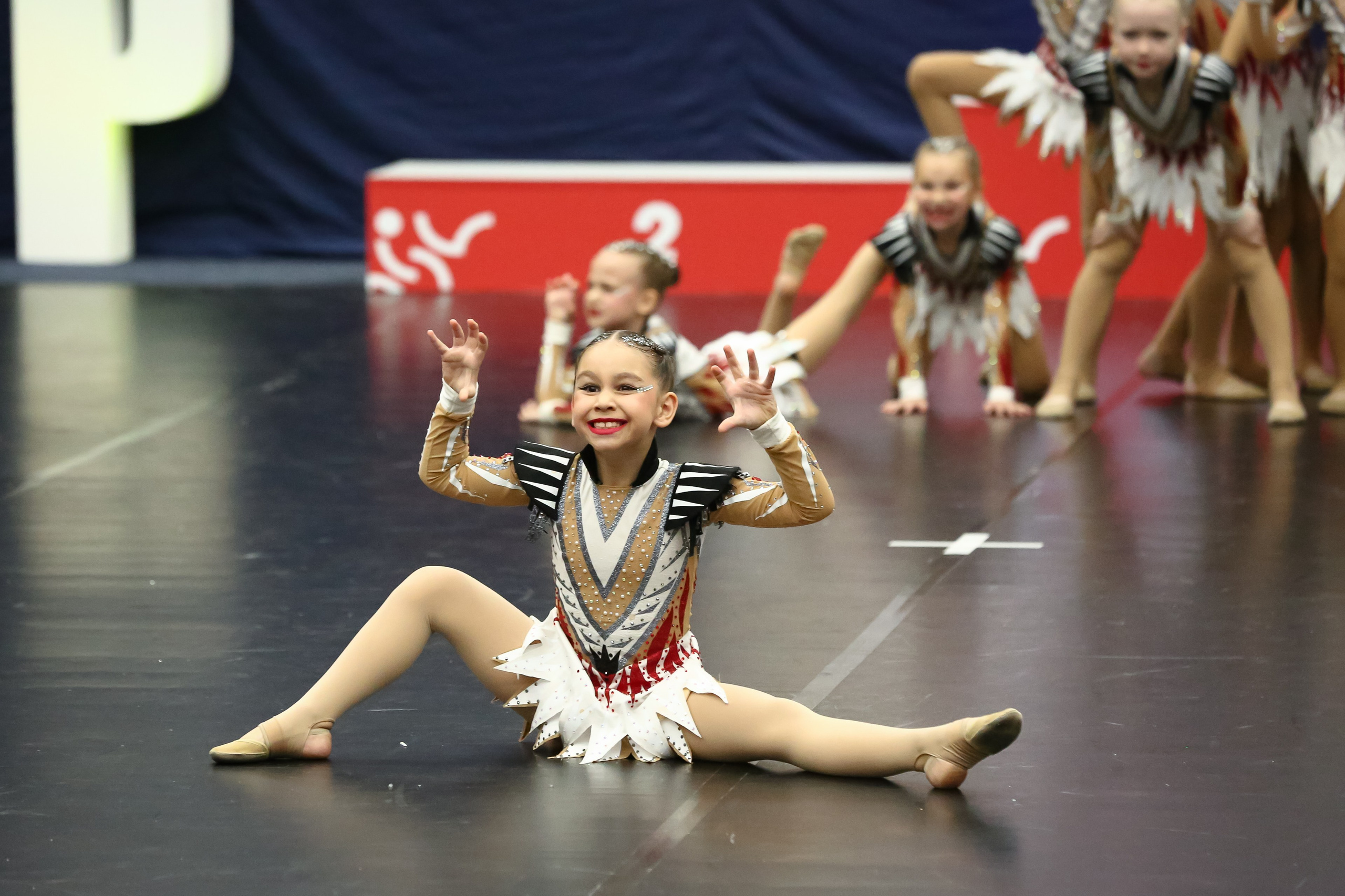 Девочка в момент выступления команды по чир спорту сидит на специальном черном покрытии, на ней красивое спортивное платье, руки она держит словно лапы тигренка с коготками. На заднем плане стоит ее команда и пьедестал для награждения.