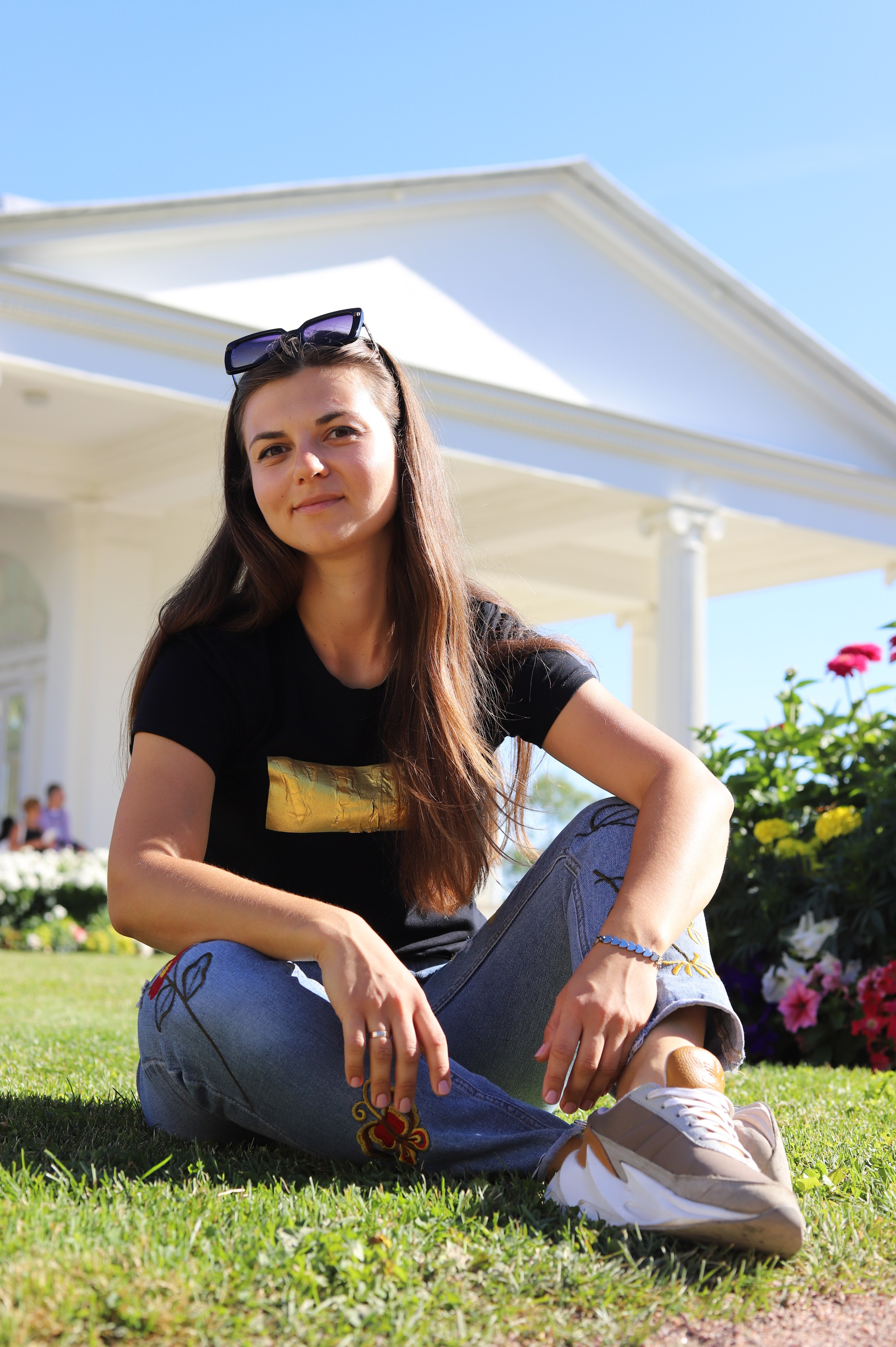 Портрет фотографа девушки на лужайке в джинсах и черной футболке, на голове девушки солнечные очки у девушки длинные волосы, девушка улыбается. девушка сидит скрестив ноги на фоне исторического здания с колоннами