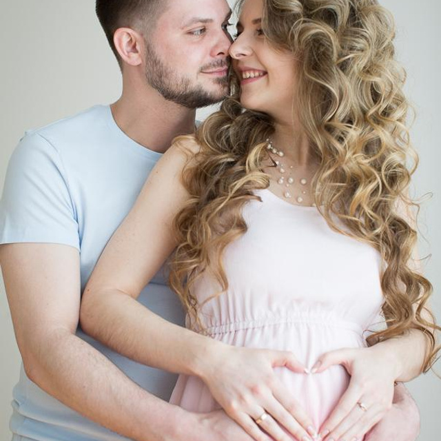 Влюбленная пара с нежностью смотрит друг на друга, из их рук сложено сердечко на животе беременной девушки