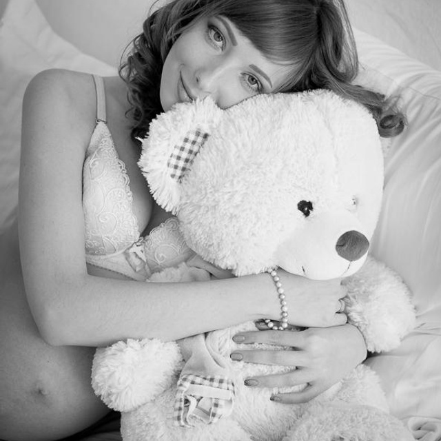 Фотосессия беременности, девушка сидит на кровати в белье, смотрит в камеру и обнимает большого белого мишку