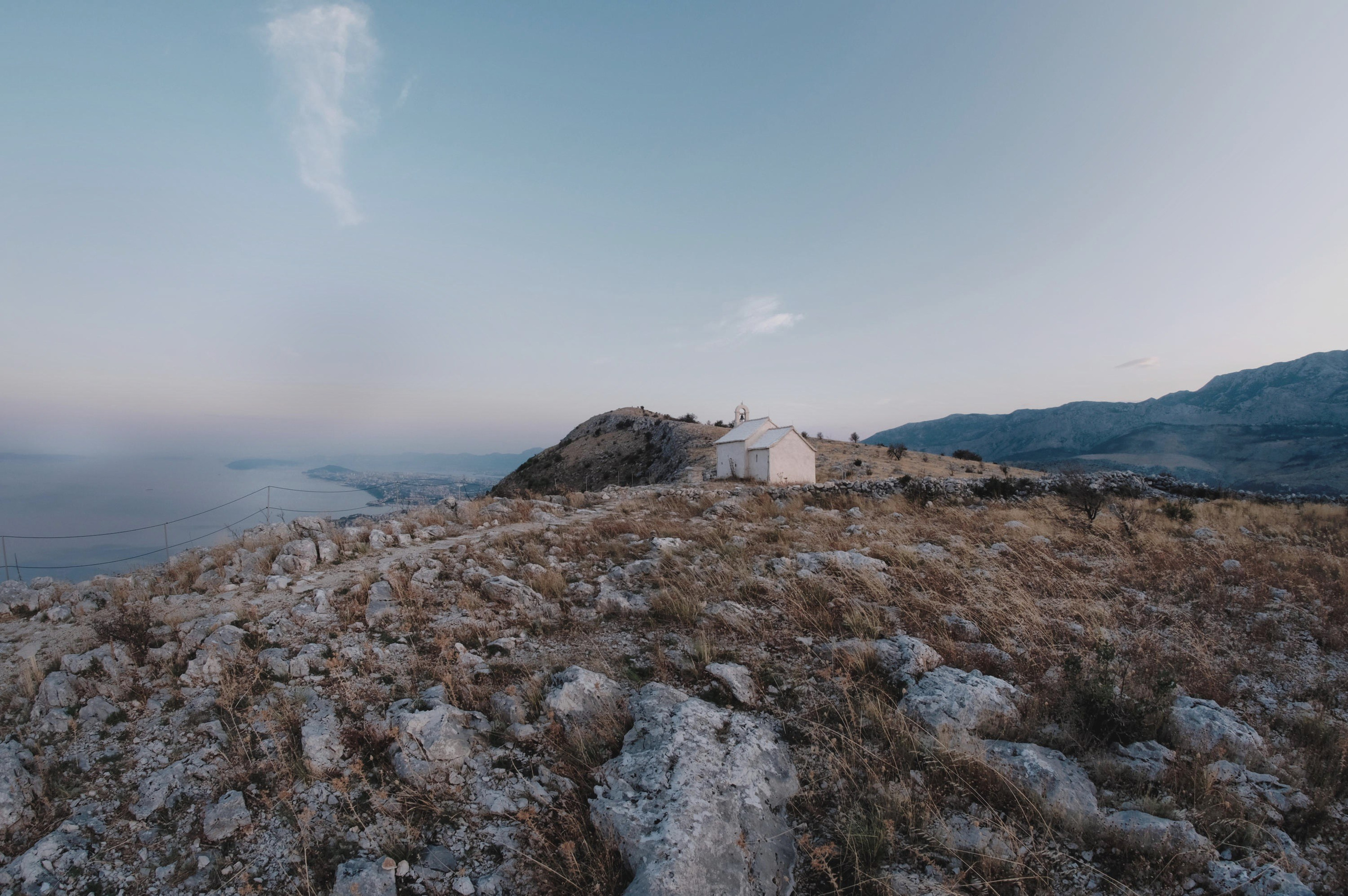 Пейзажная фотография: белая часовня на вершине скалистого холма, море и береговая линия на фоне, сухая трава и камни на переднем плане, пастельное небо