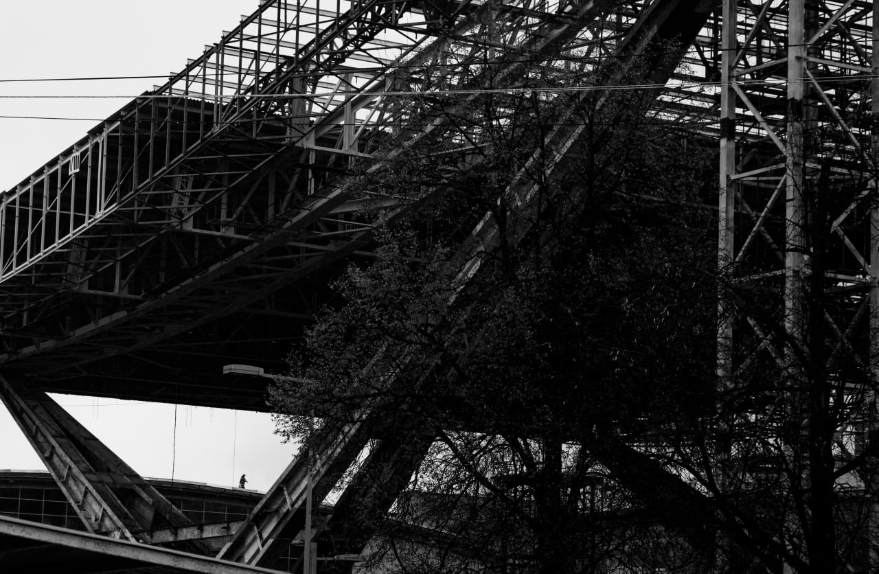 Черно-белая фотография технопустоши Павелецкой набережной. Городская фотография, отражающая концепцию археологии душ и диалога с городом в проекте Манифест Теней. Металлические конструкции, одинокая фигура вдалеке и тени деревьев создают атмосферу глубокой связи между человеком и городом. Фотография с философской глубиной, воплощающая идею "трещин в реальности", где каждый элемент становится знаком в диалоге с Москвой.