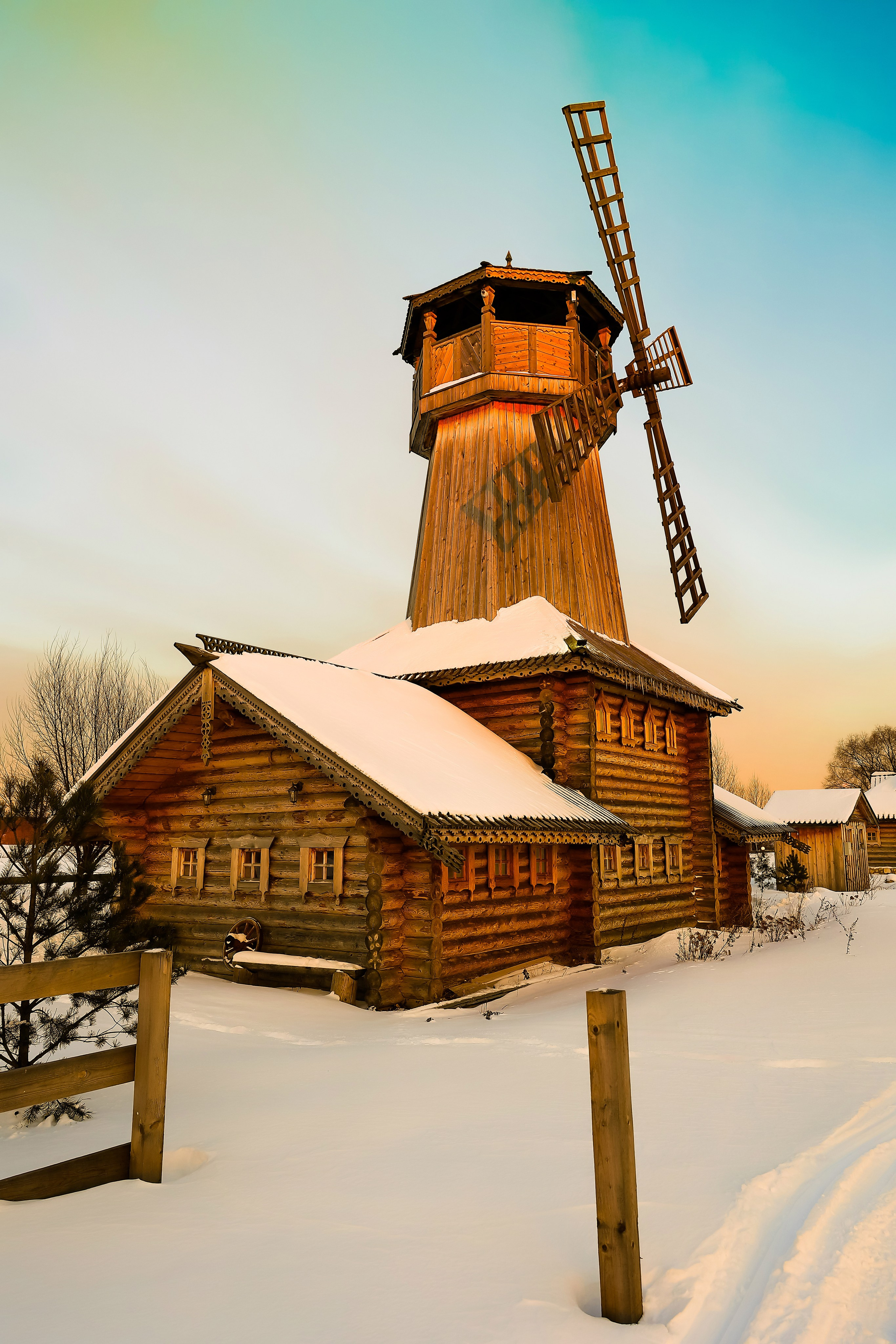 Старая бревенчатая русская мельница среди заснеженного поля, чистое небо и золотисто-оранжевый закатный свет освещают тёмные деревянные брёвна.