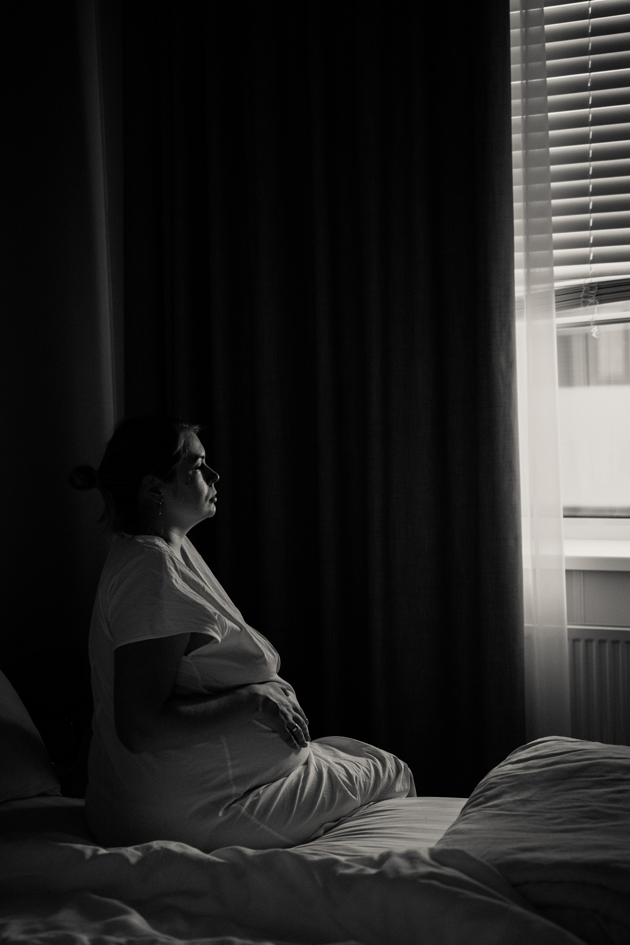 Чёрно-белая фотография беременной женщины, сидящей на кровати и смотрящей в сторону окна с жалюзи. Комната освещена мягким естественным светом через окно, создавая атмосферу покоя и ожидания. Роды, репортаж, съемка родов, роддом, первые минуты жизни.