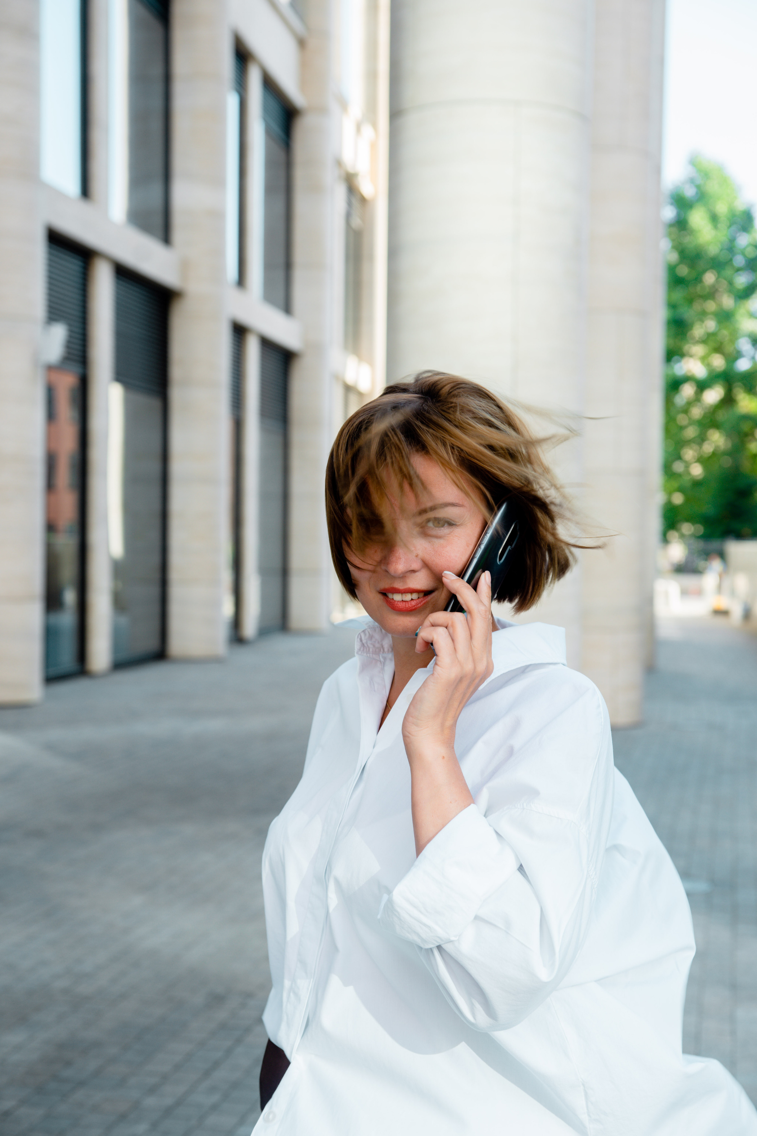 Девушка с длинными волосами, в черном костюме и сером пальто переходит дорогу. Улыбается и смотрит в камеру