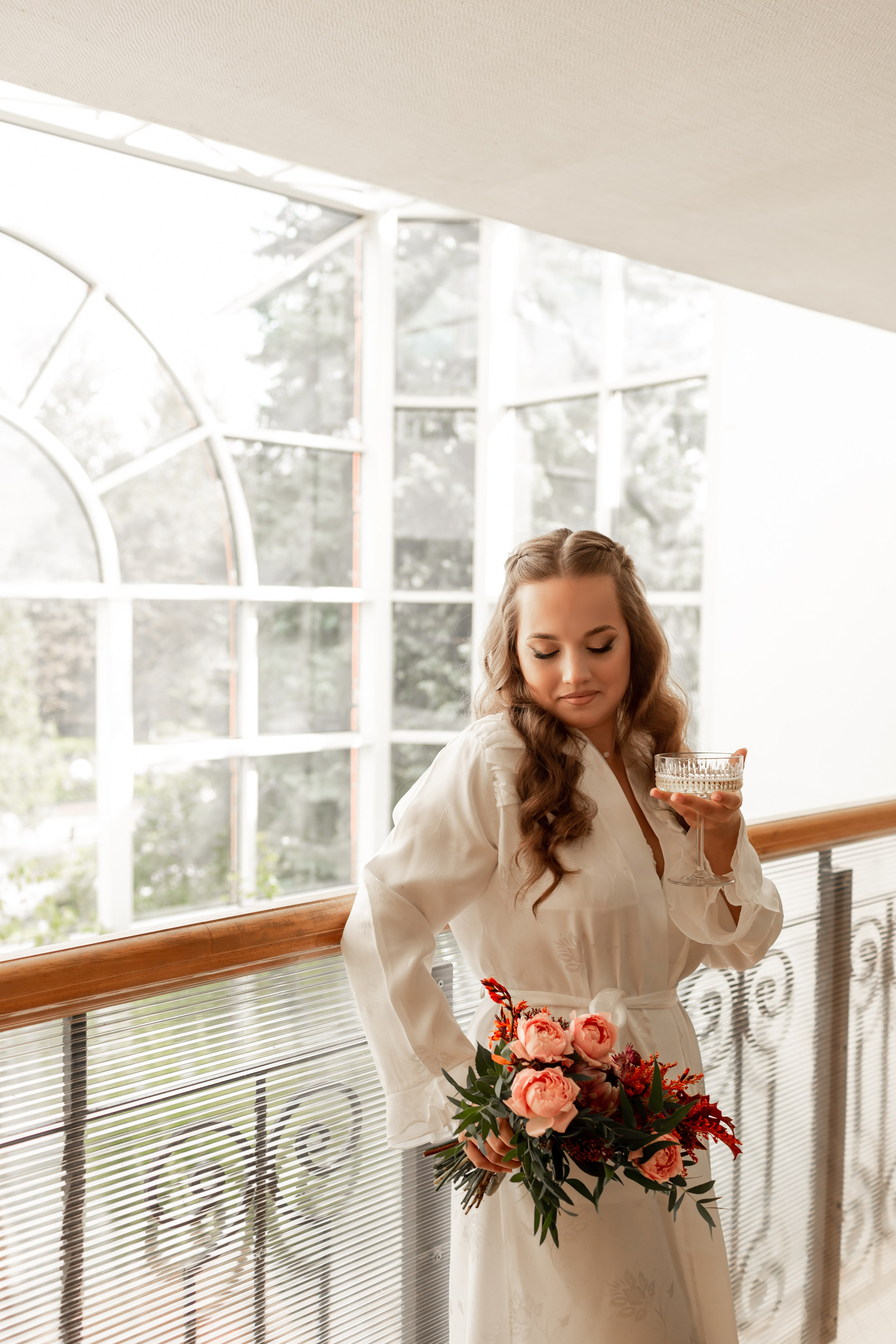 Нежная невеста в белом шелковом халате с бокалом шампанского и розовым букетом.