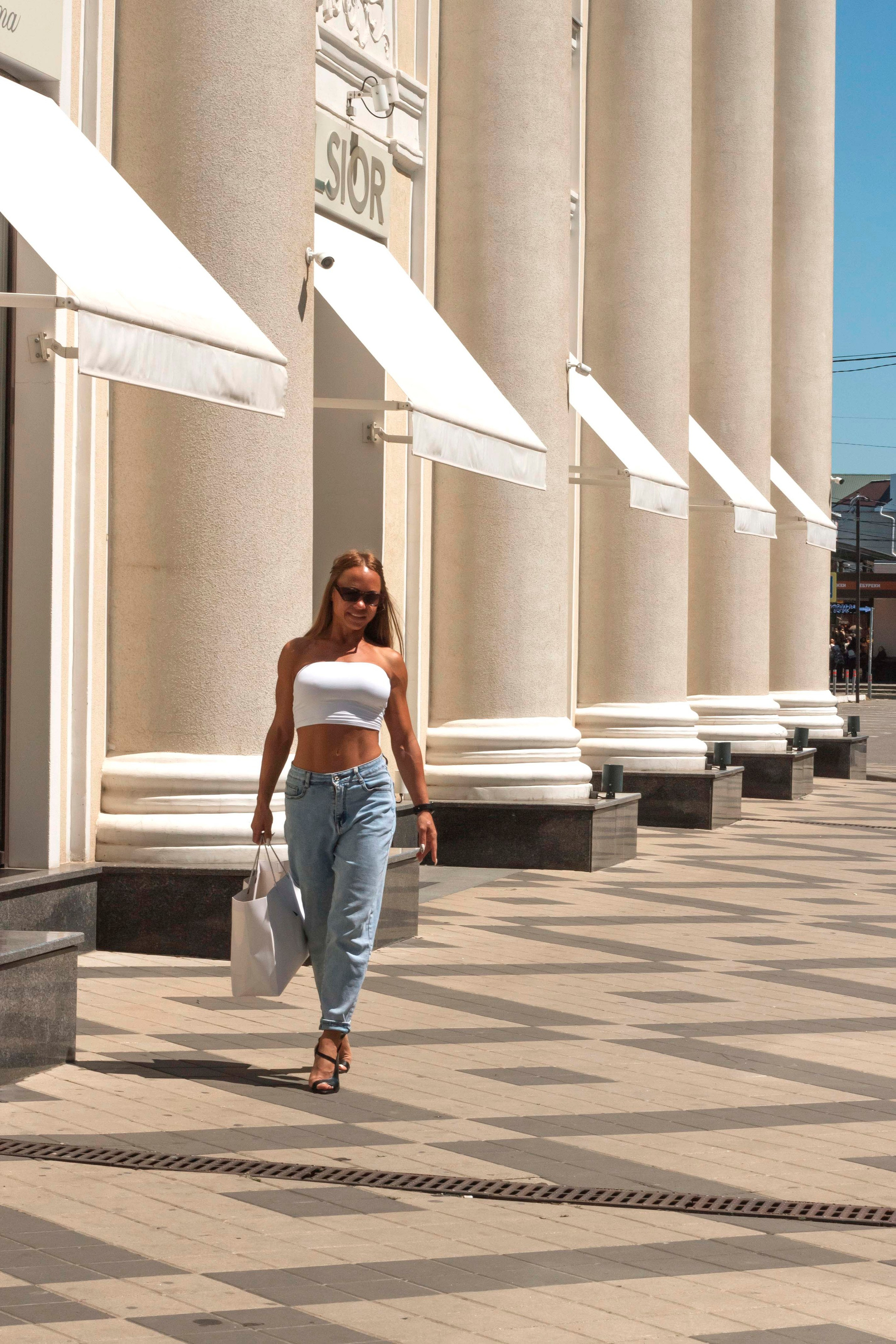 "Woman in sunglasses walks confidently through a sunny plaza lined with tall columns, white awnings, patterned pavement, and urban buildings"