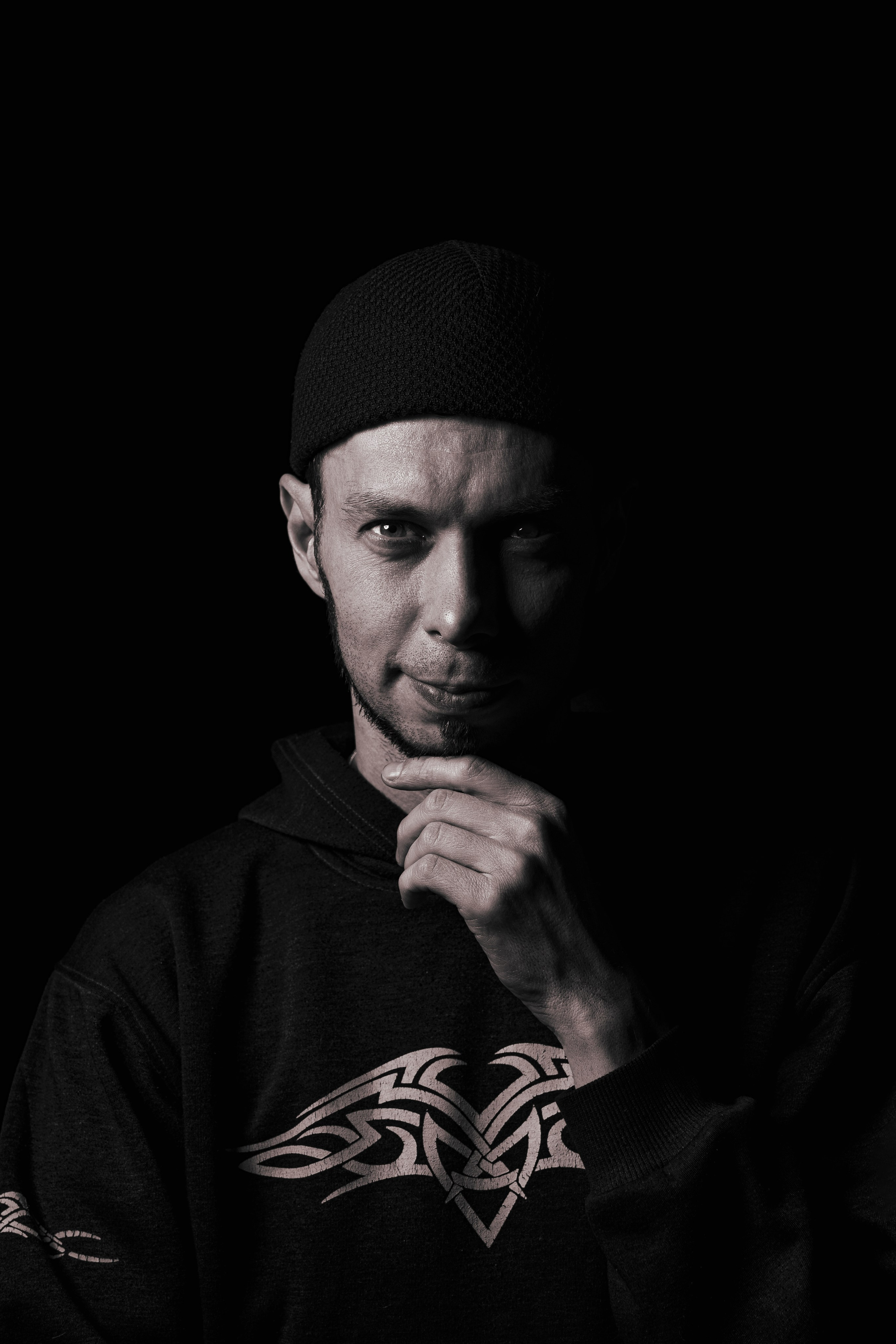 Черно-белый фотопортрет мужчины в шапке и худи с тату-орнаментом, темный фон