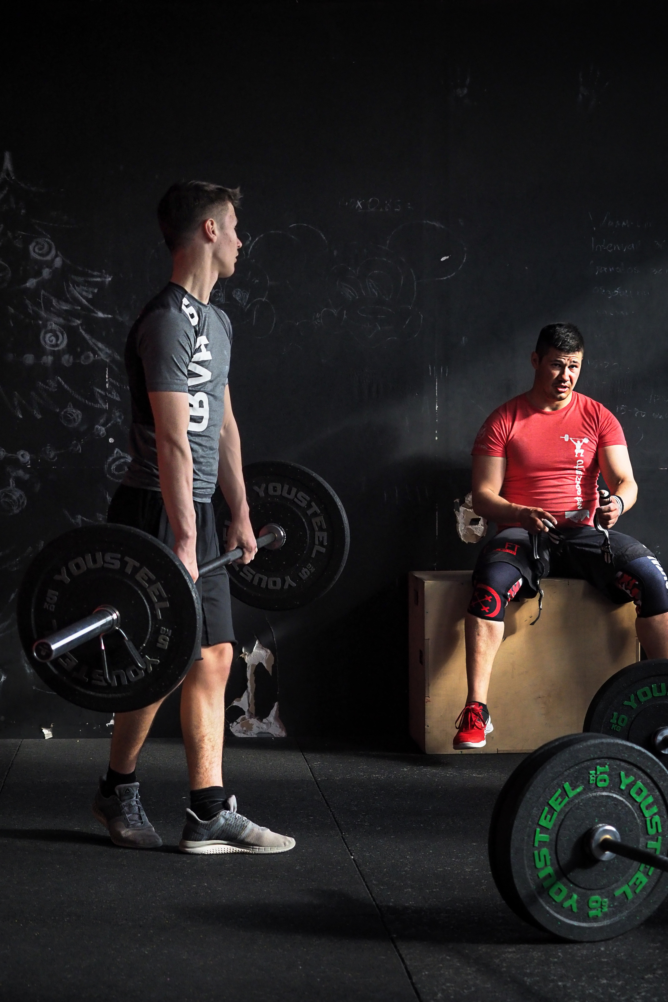 тренер сидит на тумбе и дает указание молодому спортсмену со штангой