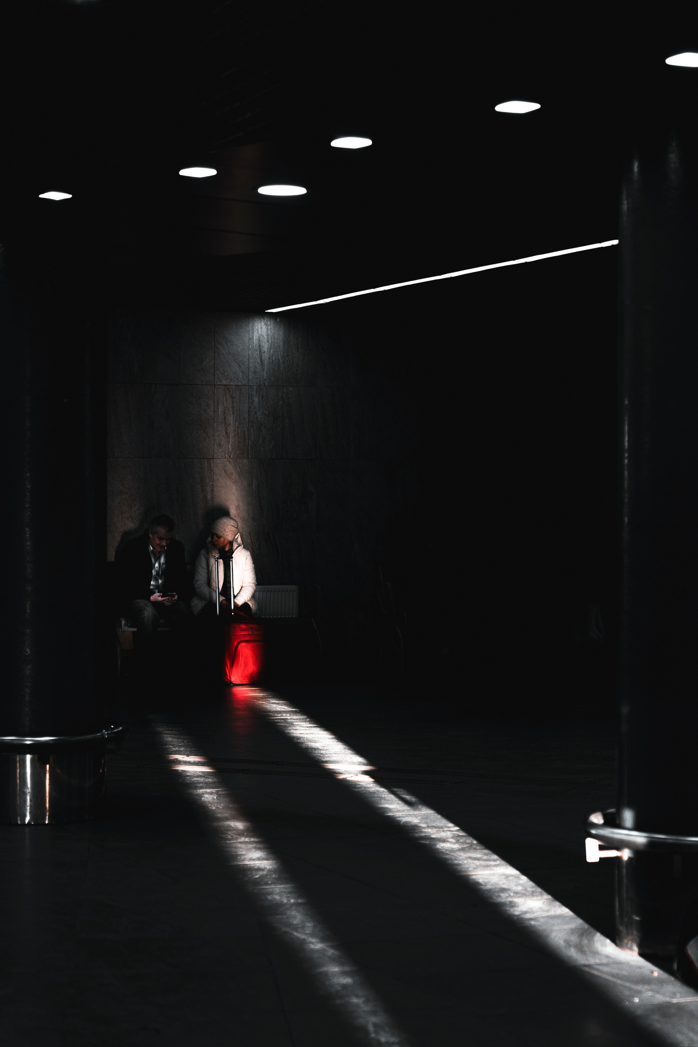 ара с красным чемоданом сидит в тёмном современном зале ожидания с длинными тенями от потолочных светильников. Стрит-фотография Мальцева Евгения.