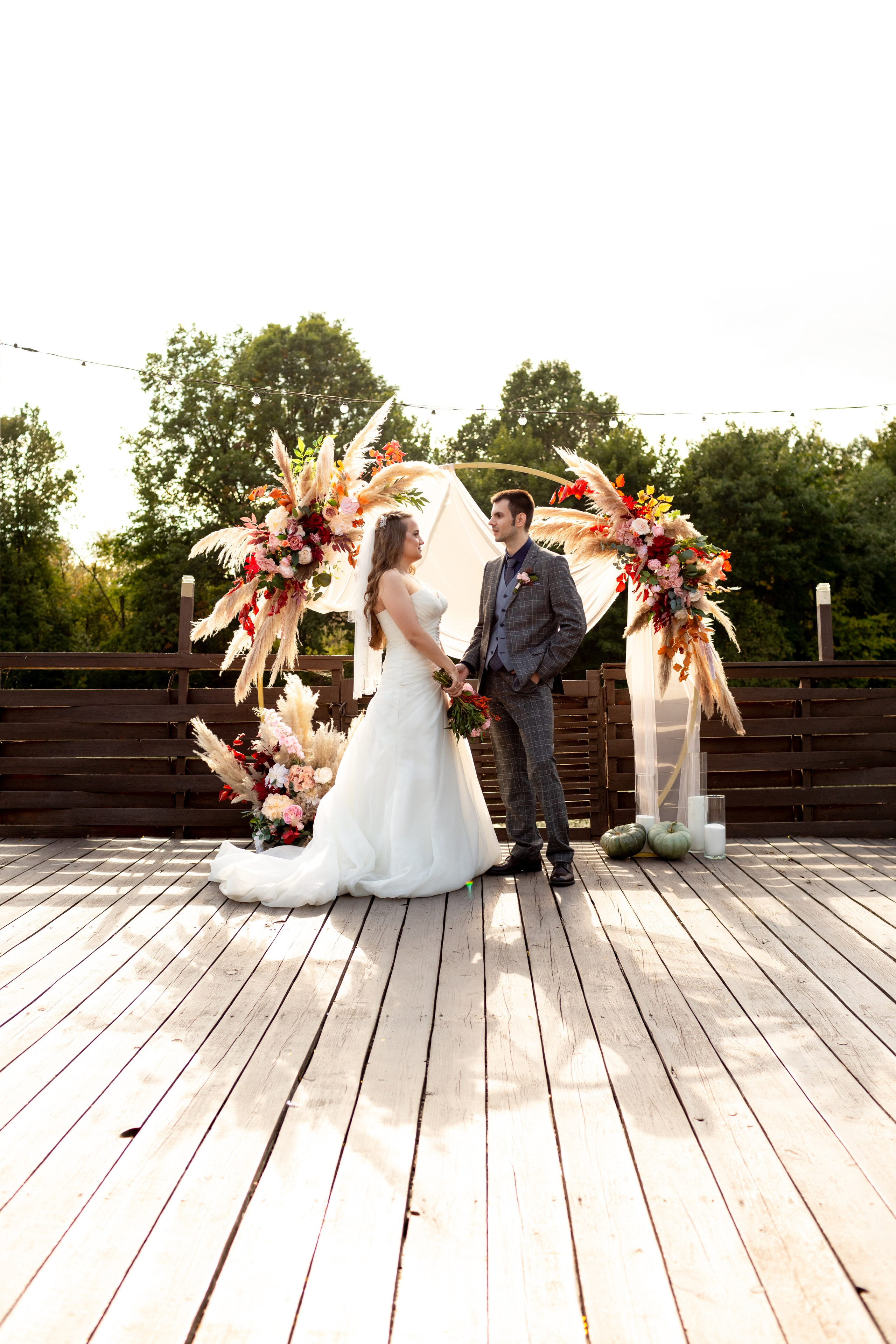 Выездная регистрация: молодожены держат друг друга за руки у арки с цветочными украшениями на берегу озера.