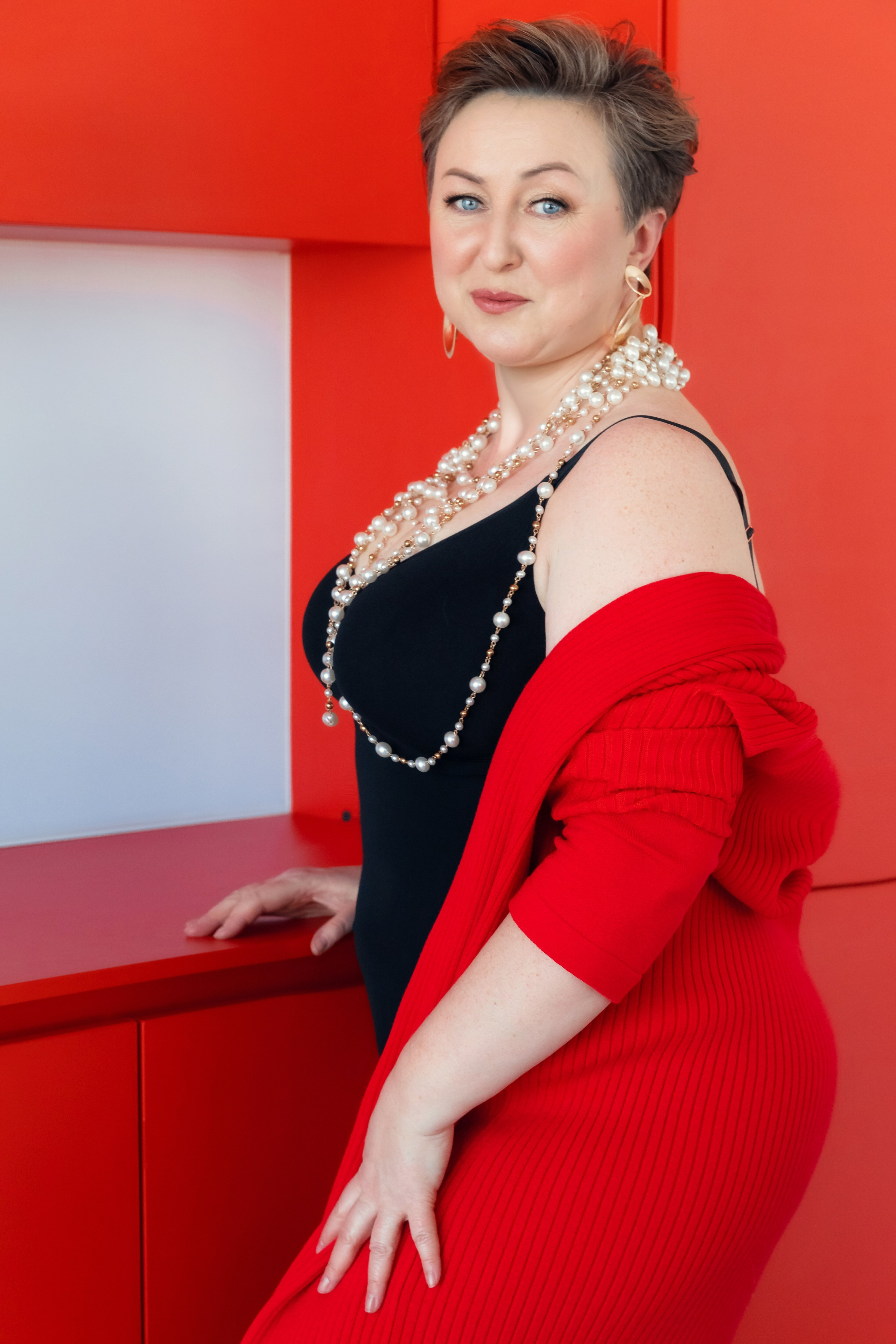 Женщина в черном платье и красном кардигане лежит на красной глянцевой поверхности, стильная фотосессия с акцентом на украшениях и образе