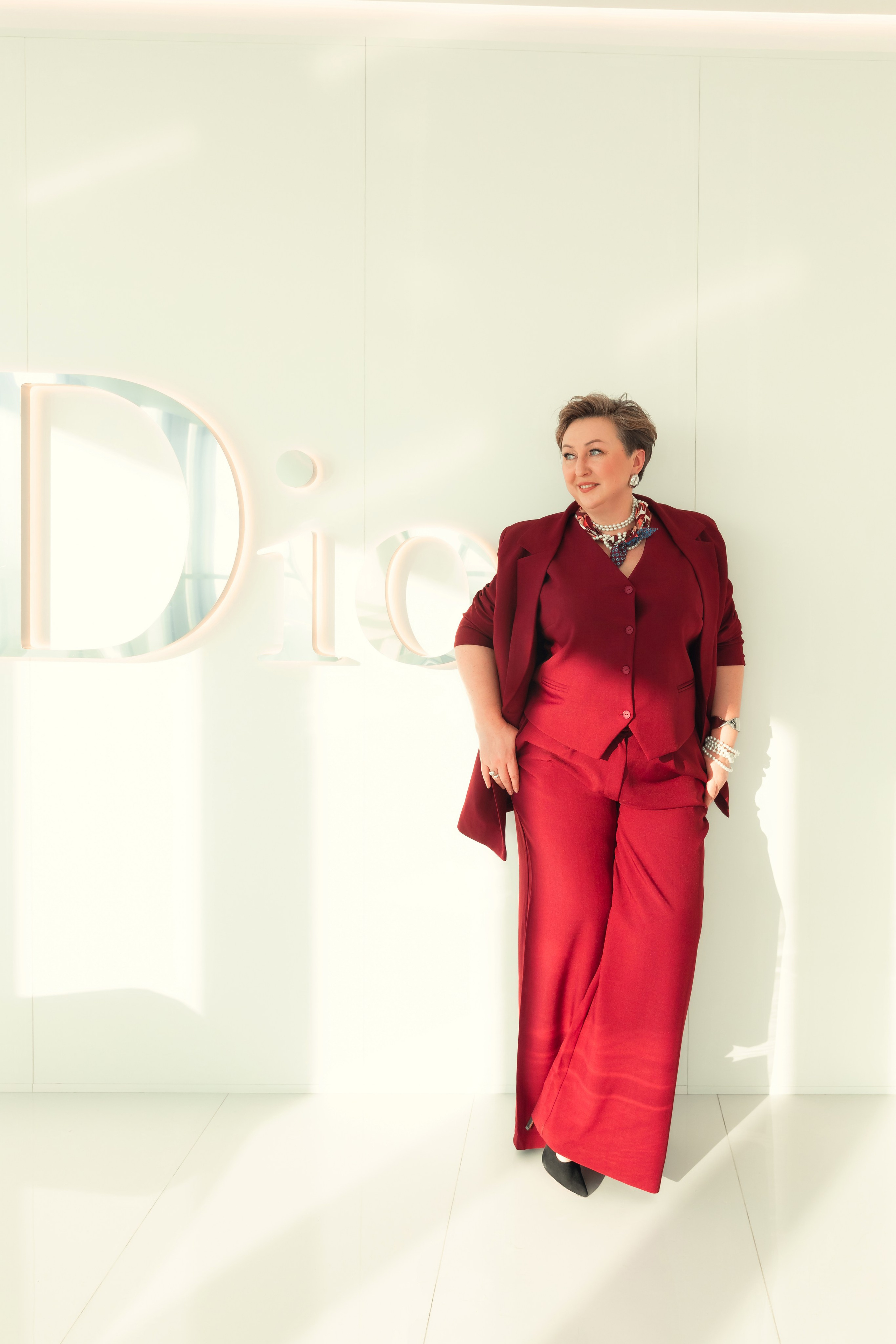 Женщина в бордовом костюме и украшениях позирует на светлом фоне, деловой и элегантный стиль.