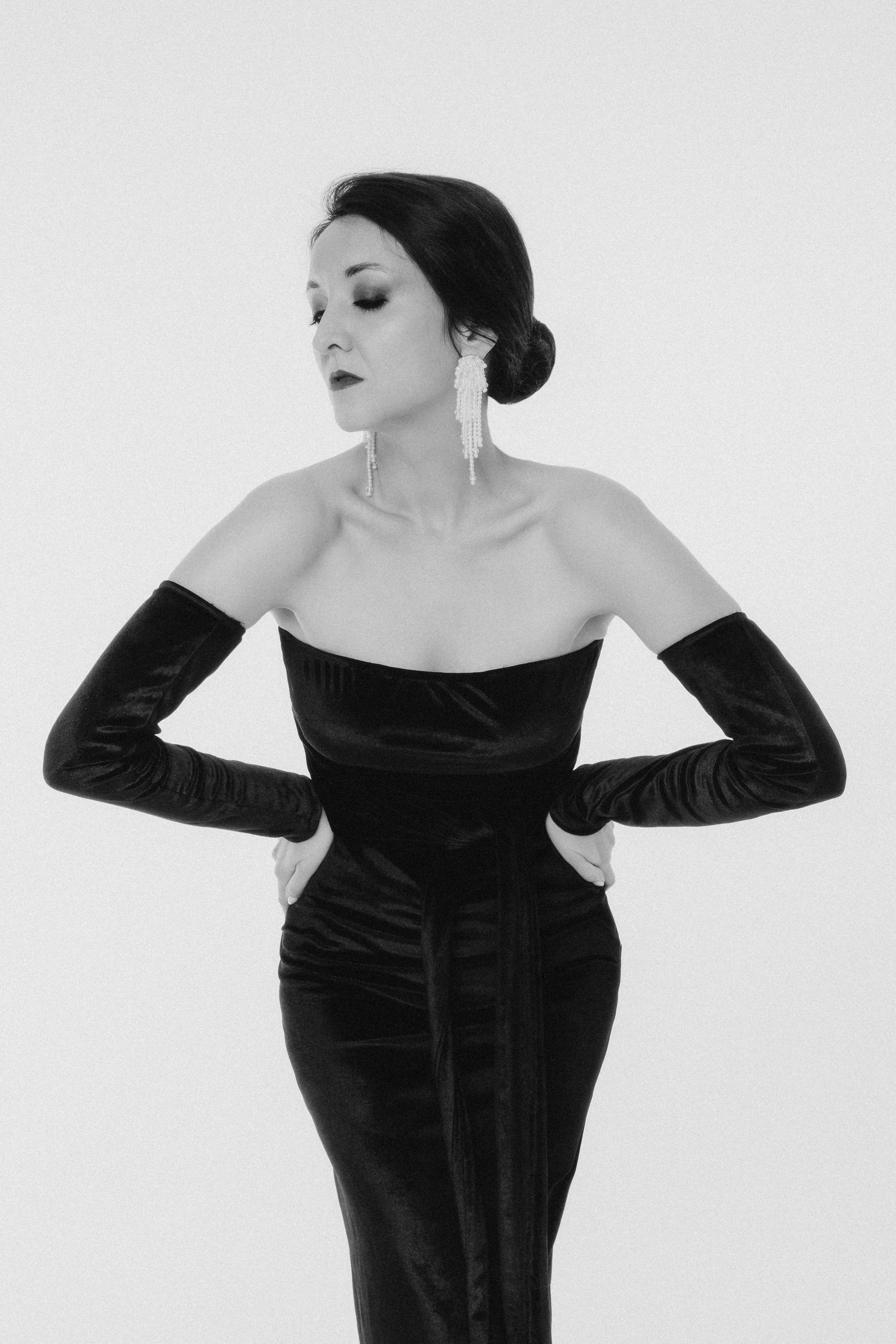 Женщина в длинном черном платье с открытыми плечами и длинными перчатками, позирует на белом фоне. Женщина в черном вечернем платье элегантно позирует, светлый нейтральный фон, черно-белое фото.