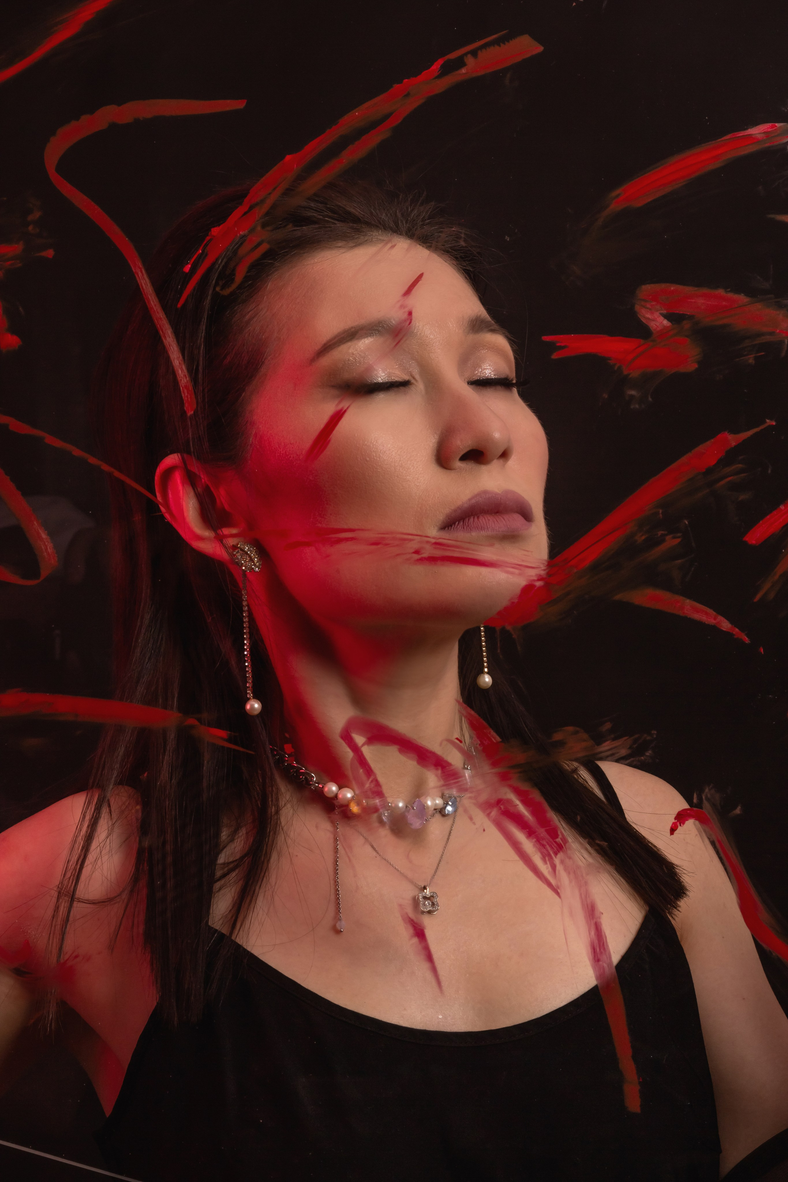 "studio photo A woman with closed eyes and pearl jewelry is framed by vivid red paint-like streaks against black, creating a dramatic, haunting portrait.