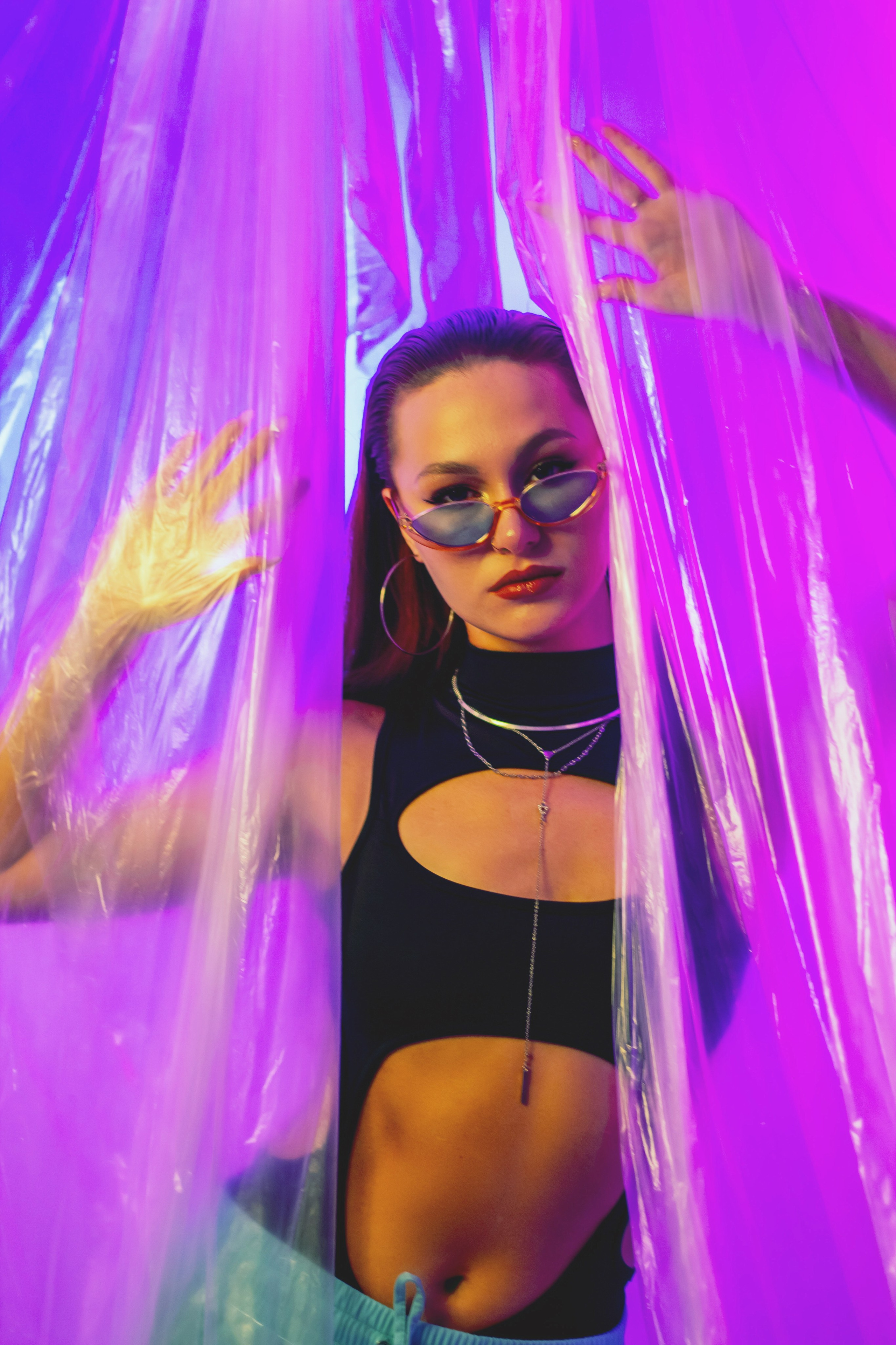 "Woman in sunglasses poses behind translucent plastic strips under vivid purple and pink lighting, creating a bold, futuristic, mysterious mood".