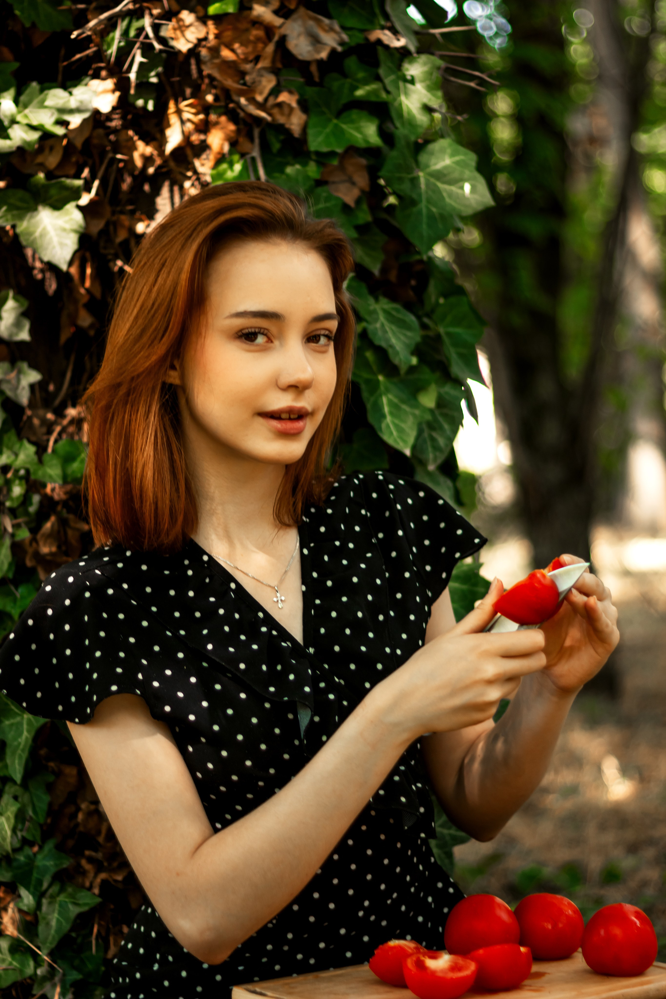 "Young woman in a black polka-dot dress slices a red apple in a leafy forest, with more apples nearby; the scene feels warm and peaceful".