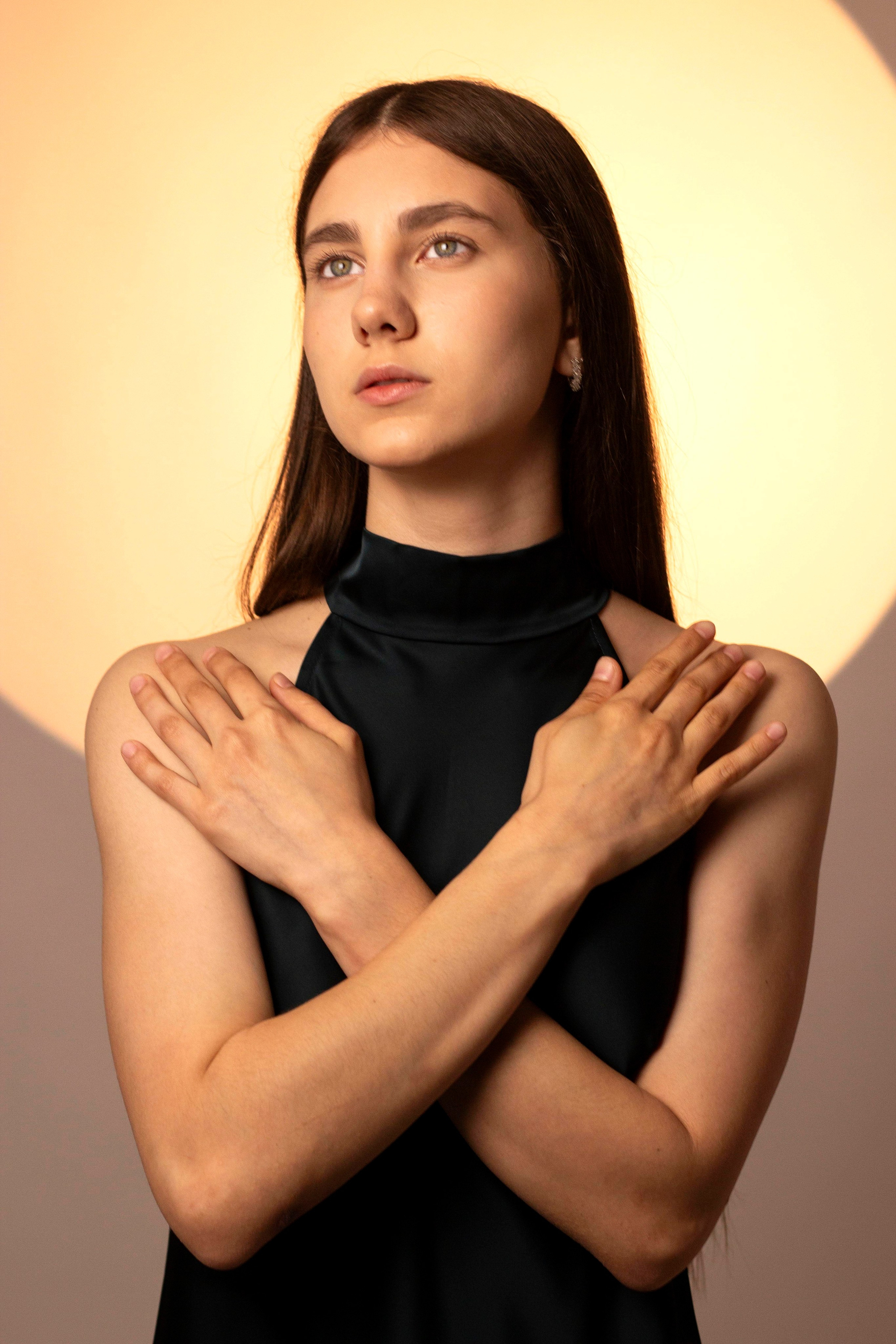 "Portrait of a woman in a black sleeveless top crossing her arms in a self-embrace, gazing upward with a calm, reflective expression".