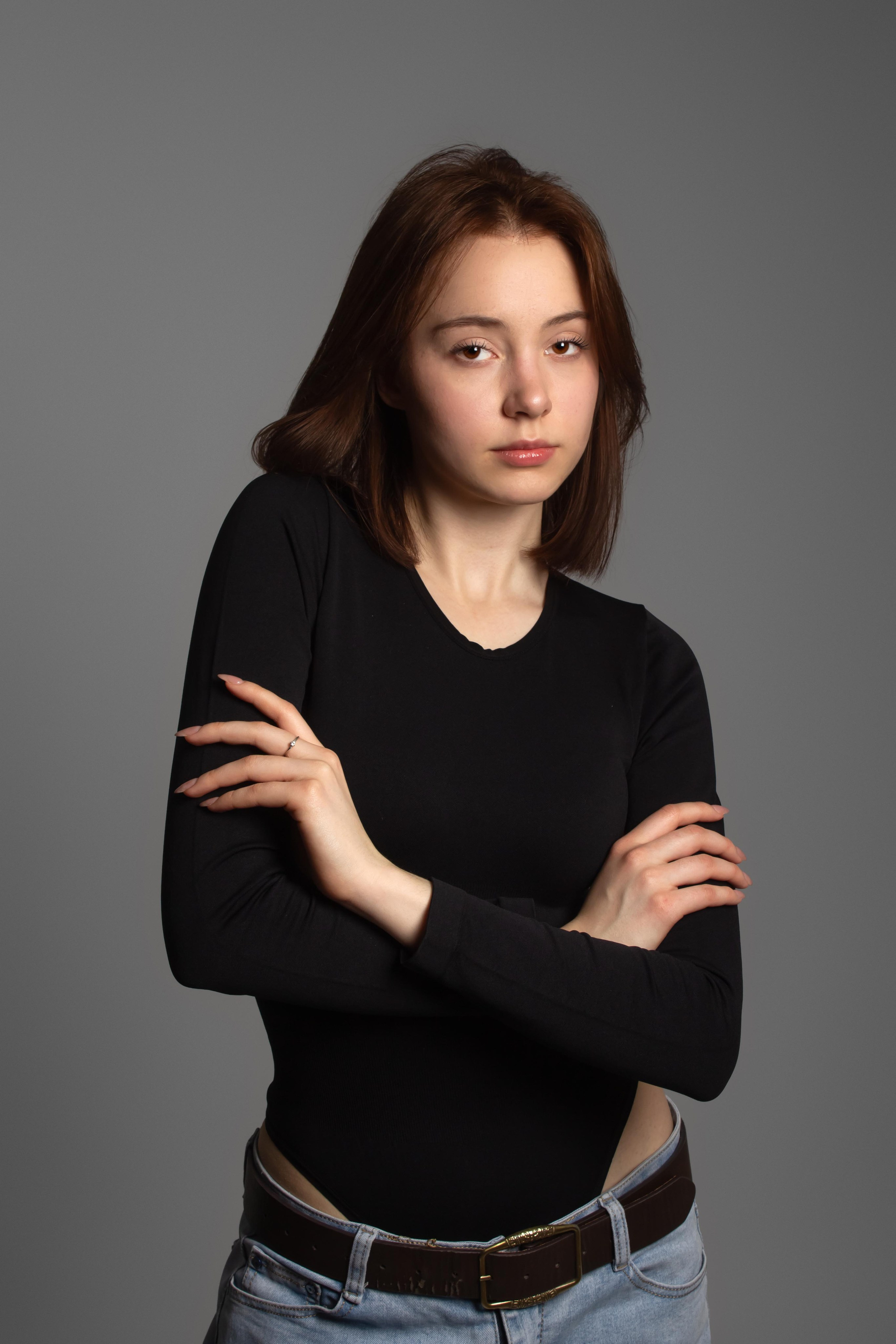 "Portrait of a young woman with brown shoulder-length hair in a black top and jeans, arms crossed, gazing calmly at the camera against gray".