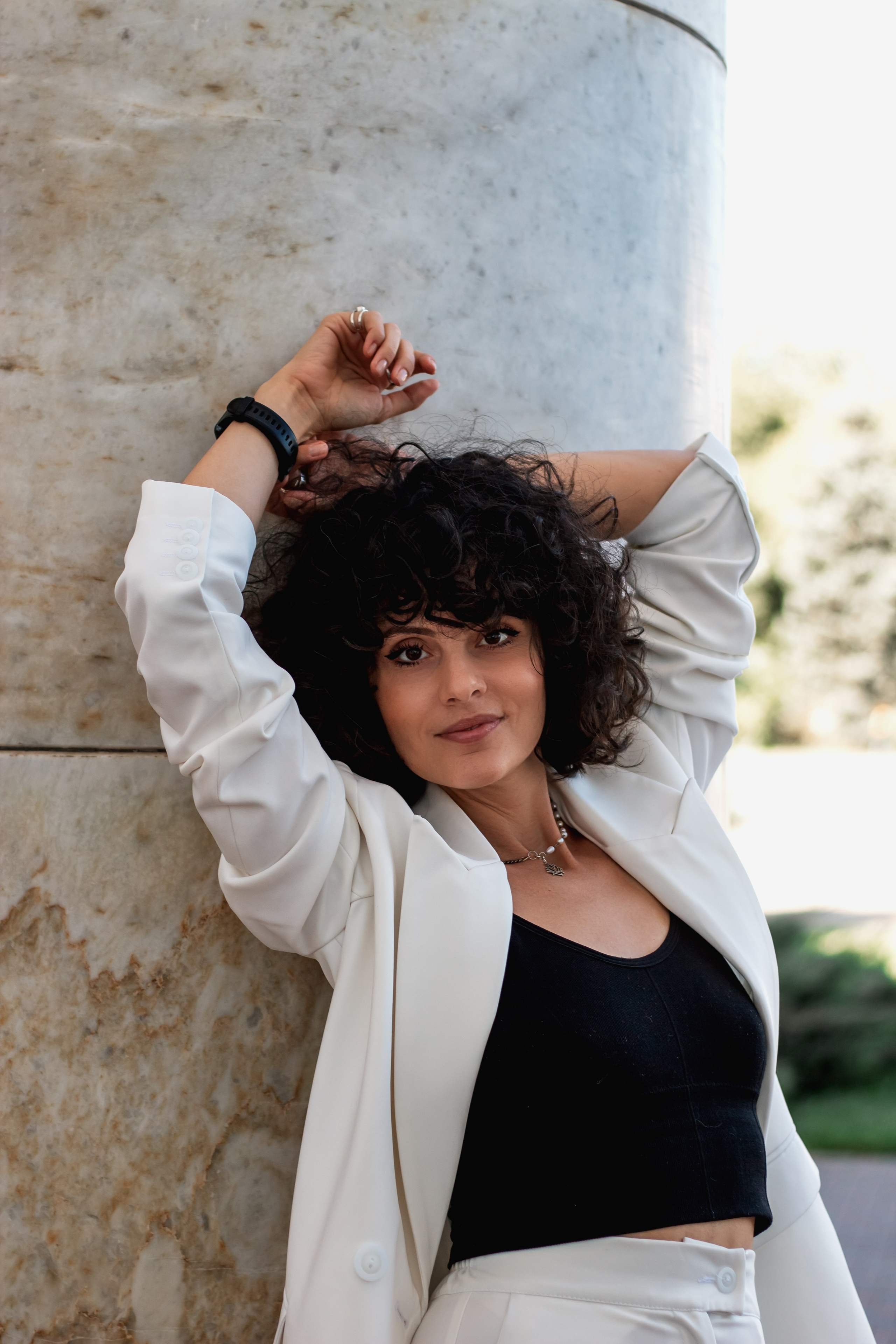 "Curly-haired woman in a white blazer and black top leans against a stone column, gazing at the camera with a relaxed, confident expression".