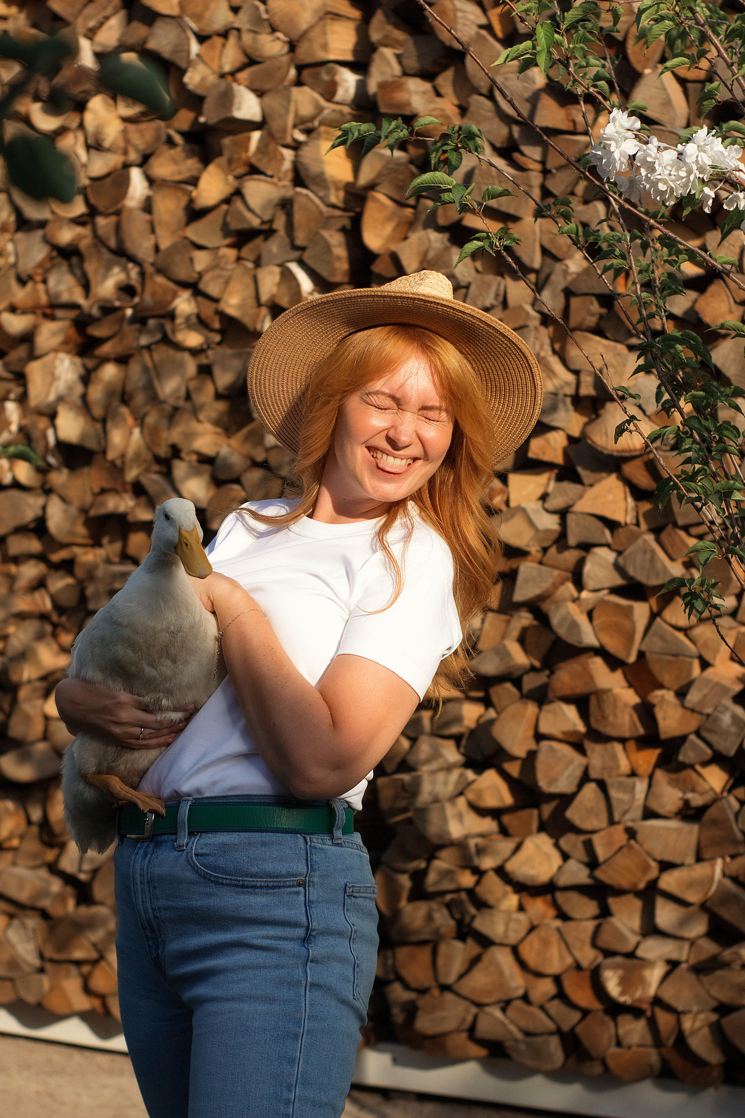 Рыжая девушка в соломенной шляпе на фоне дровника держит гуся на руках и мило улыбается.