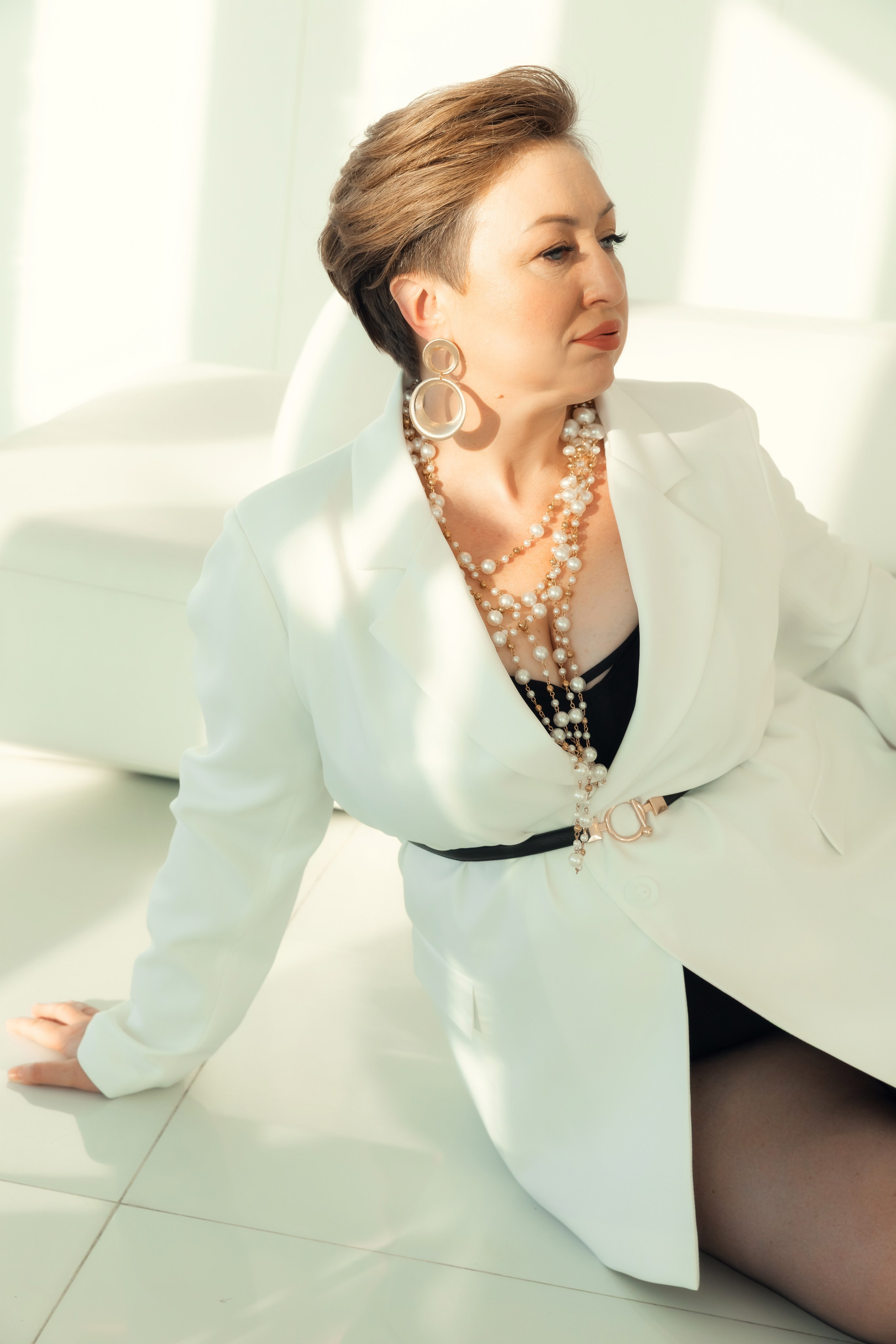 Женщина в белом пиджаке и массивных серьгах сидит на полу в светлом интерьере с солнечным светом, современный женственный портрет. Женщина с короткой стрижкой позирует в современном интерьере, стильные аксессуары и спокойное выражение лица.