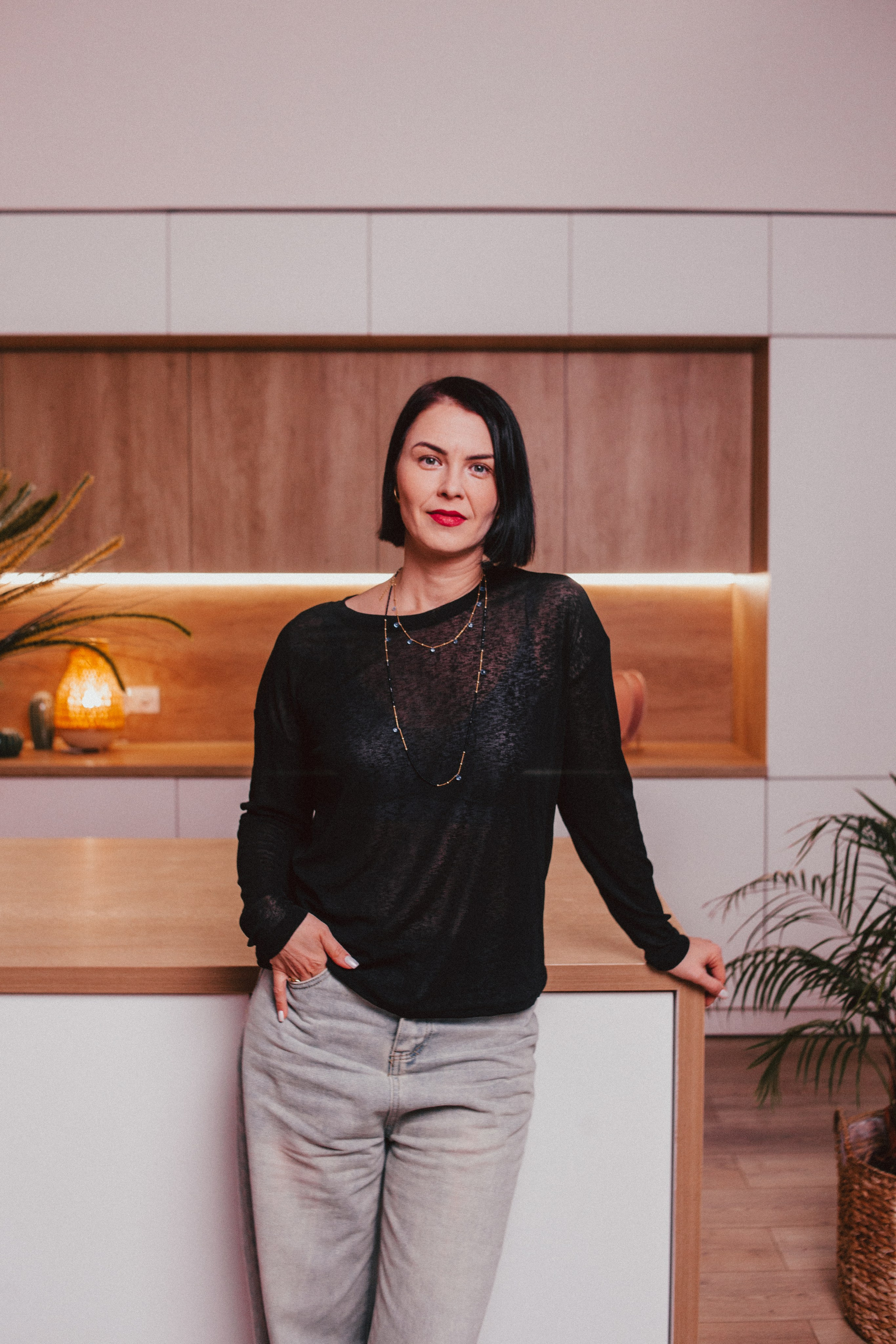 Женщина в кофте и джинсах позирует в хорошо обставленной комнате.