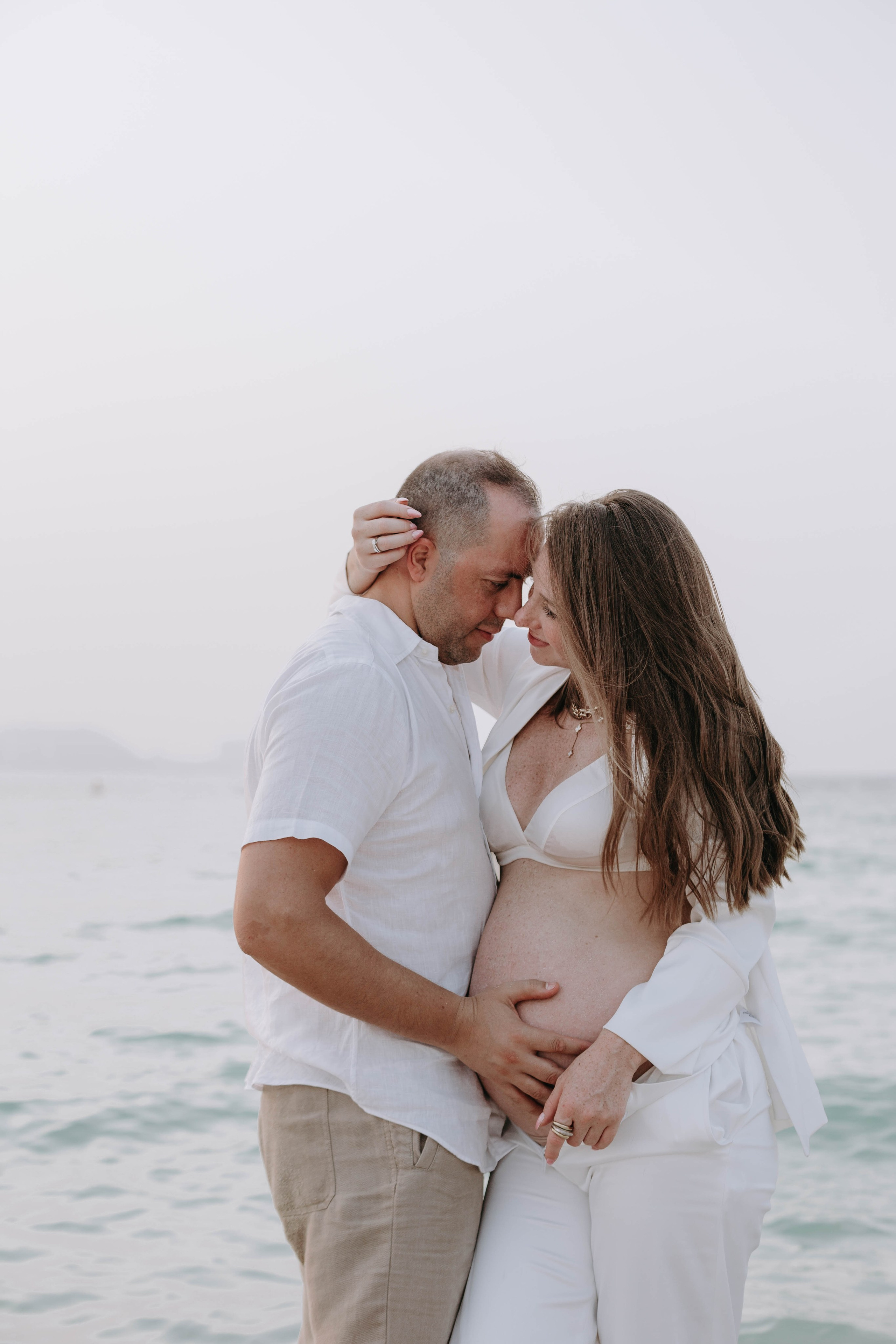 A pregnant woman and her partner embrace on the beach, foreheads touching and hands on her belly, sharing a loving moment by the sea