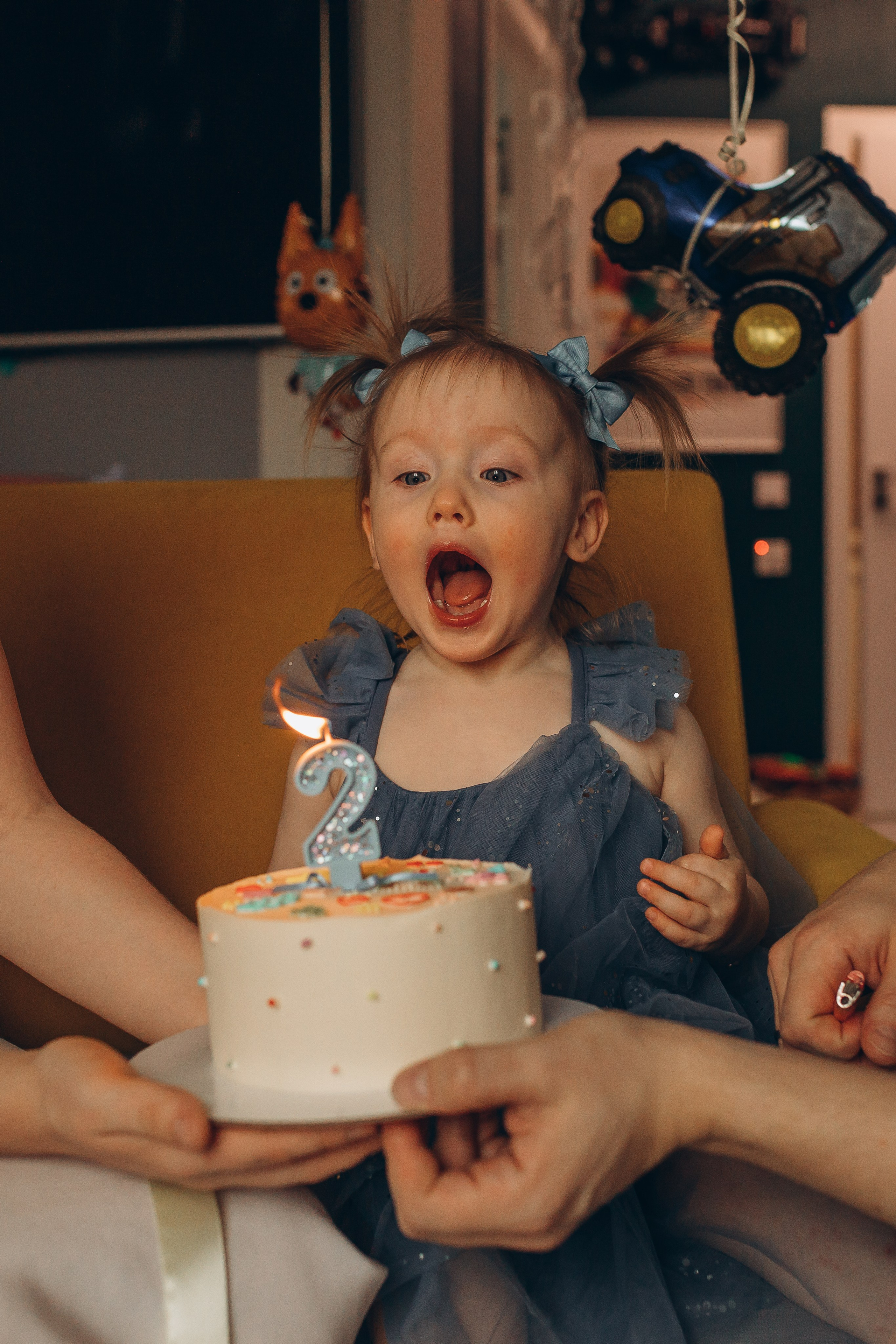 Маленькая счастливая девочка в синем платье с голубыми бантиками набирает воздух чтобы задуть свечу на своем торте в день рождения. Фотограф Маруся Самылкина в Санкт-Петербурге.