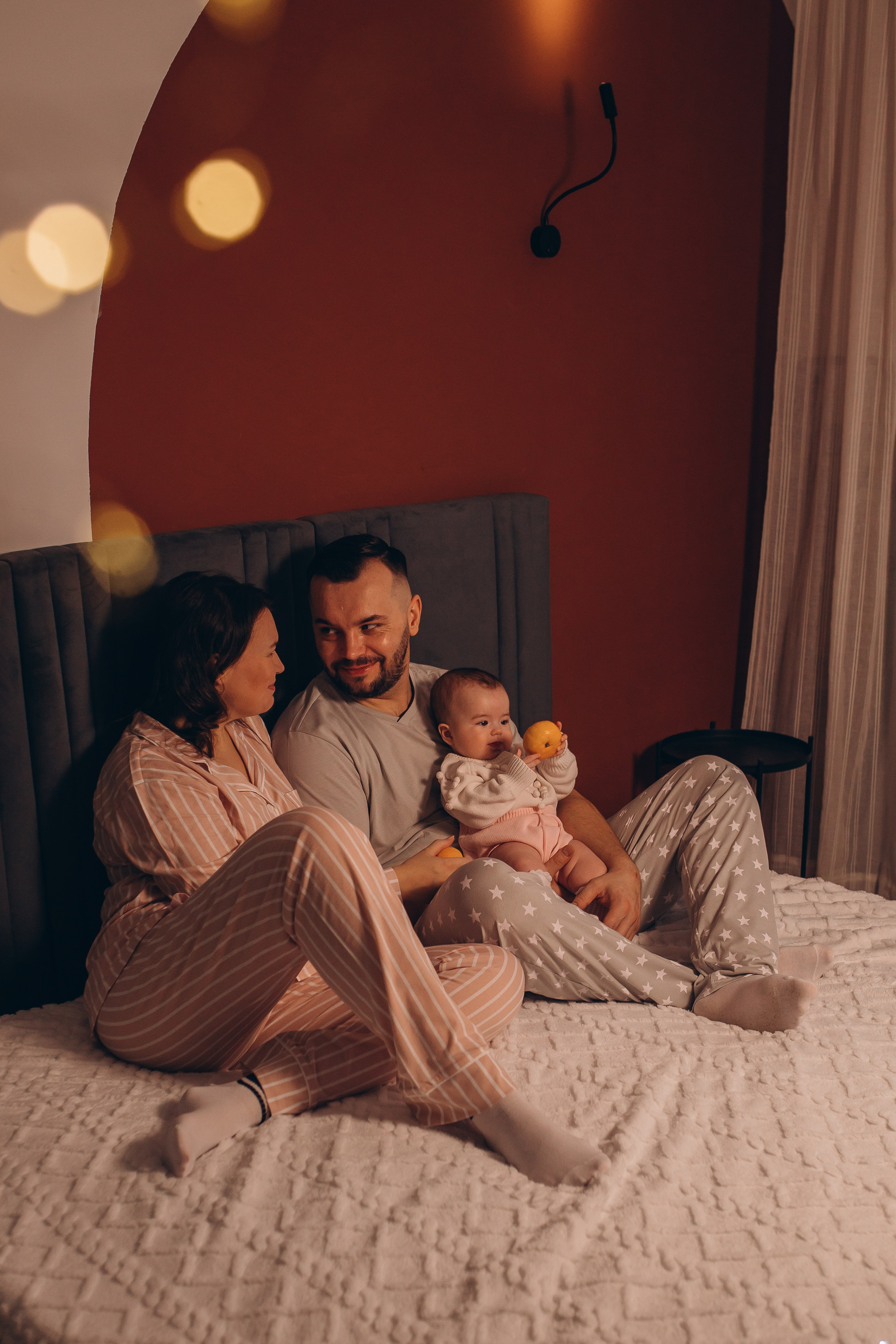 Счастливая семья: мама и папа держат малышку на руках на кровати на фоне контрастной стены. Фотосессия для малышей и новорожденных в Санкт-Петербурге, фотограф Маруся Самылкина.