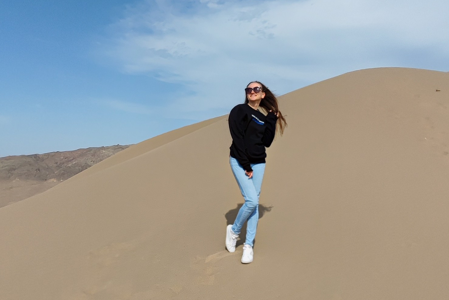 Человек позирует на фоне бирюзового озера Иссык в районе Алматы и гор. Он стоит на большом камне, одет в ярко-зеленую толстовку, черные штаны и светлую обувь, а также оранжевую панаму. В руках держит очки. За ним виднеются деревья, лесистые склоны и горные вершины под облачным небом. Атмосфера спокойная и природная.