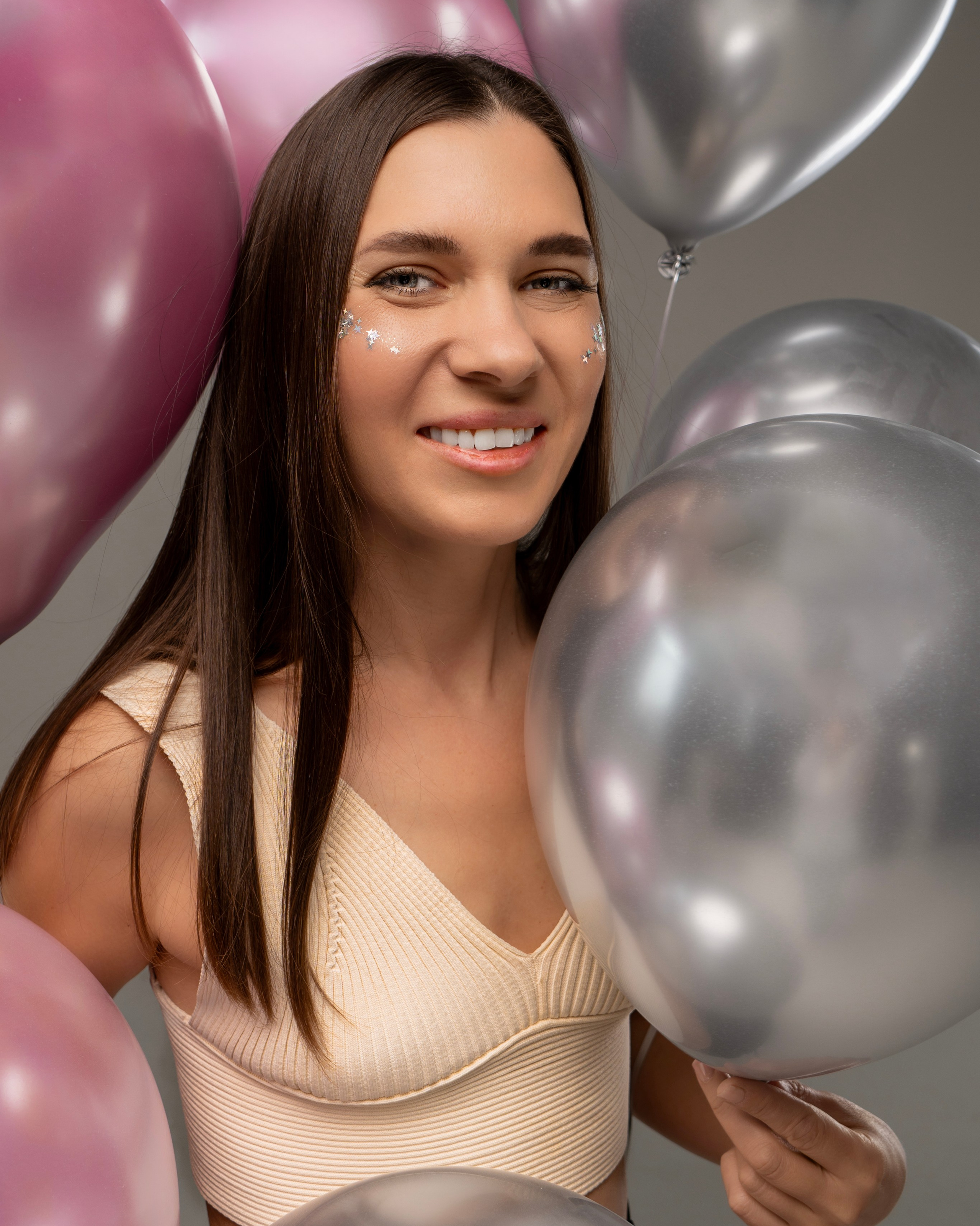 Улыбающаяся девушка с длинными тёмными волосами в светлом топе, окружённая серебристыми и розовыми воздушными шарами, с блестками в виде звёзд на щеках.