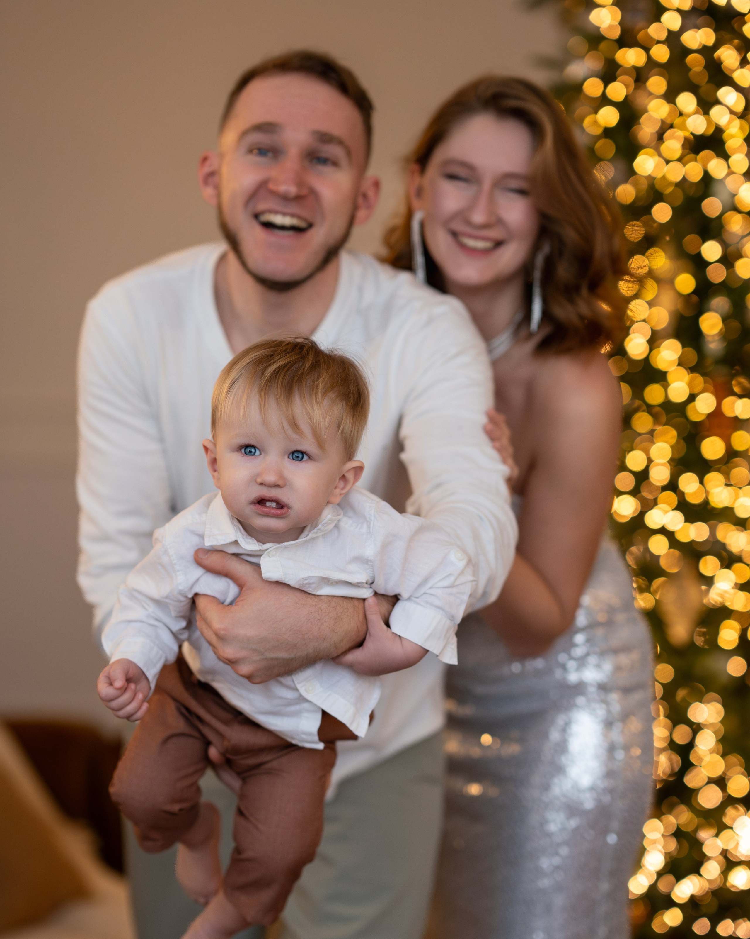 Счастливая семья с малышом на руках на фоне новогодней ёлки с огоньками: мужчина в белой рубашке держит ребёнка, женщина в блестящем платье улыбается рядом.