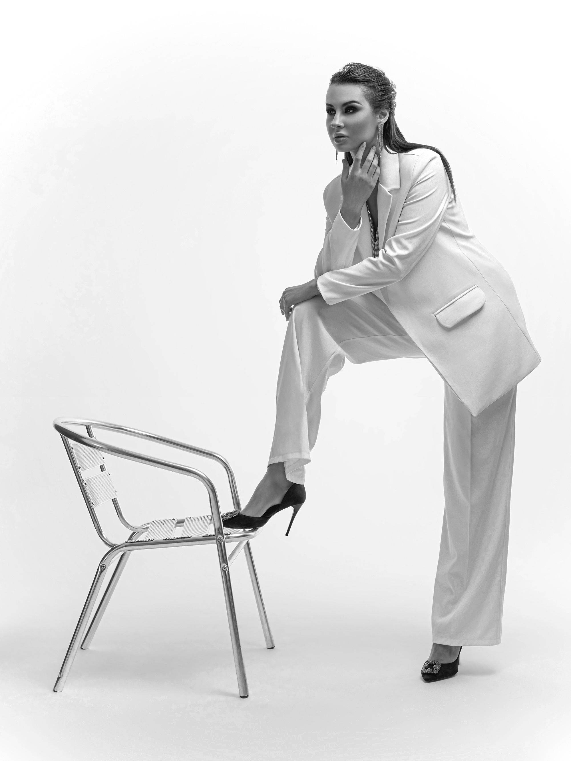 Девушка в белом костюме и в черных туфлях поставила ногу на металлический стул