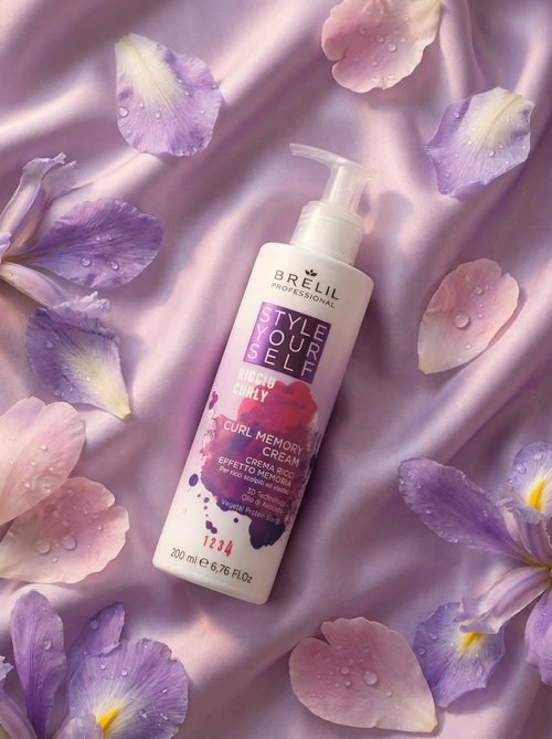 Продукт лежит на шелковой ткани лавандового цвета.  Вокруг флакона разбросаны лепестки ирисов и пионов, а также несколько капель росы которые выглядят как застывшие капли парфюма. 